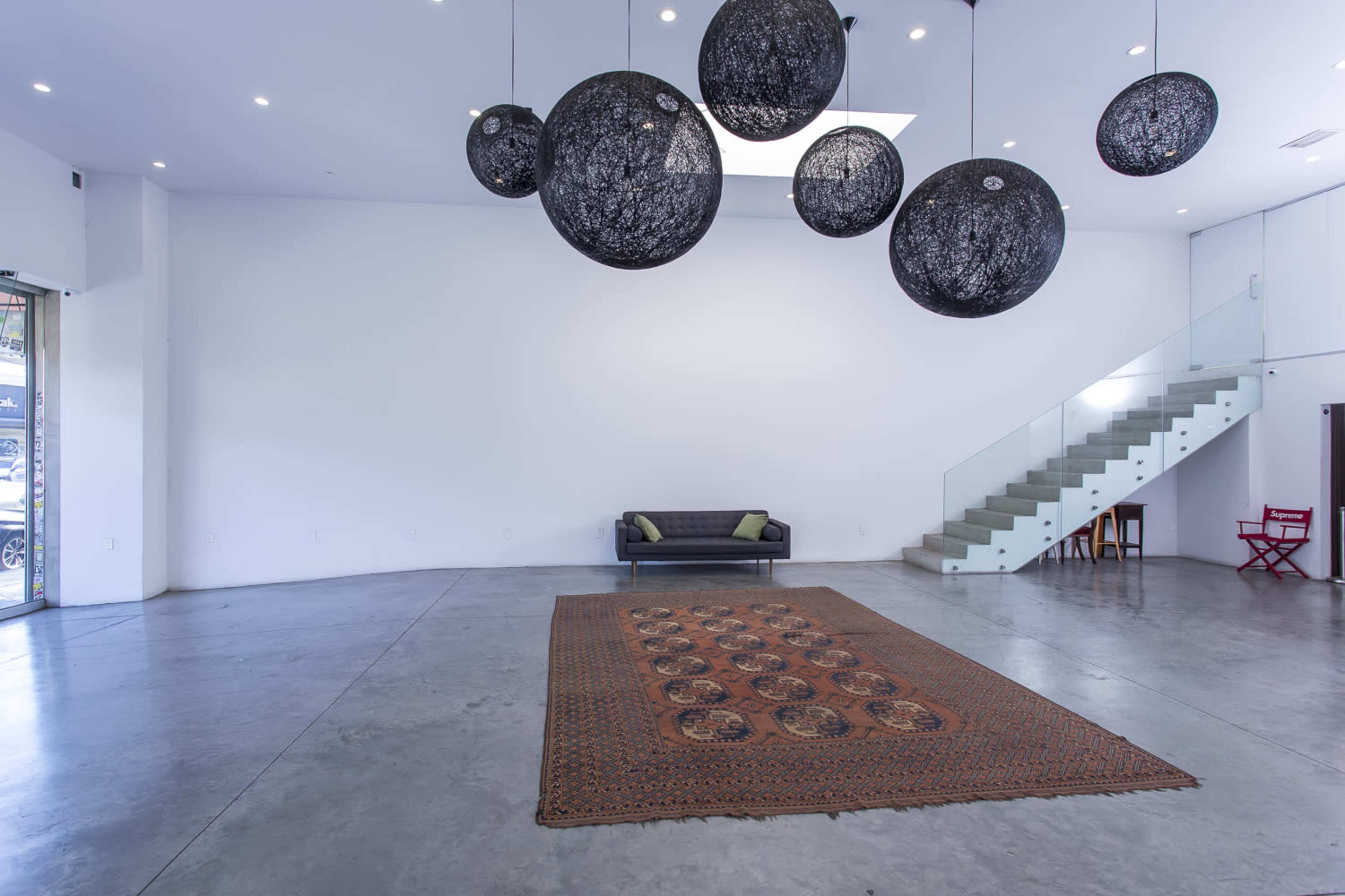 The image shows a spacious, modern interior with a large, textured rug on the concrete floor, a black sofa, and hanging round light fixtures above, with a glass staircase in the background.