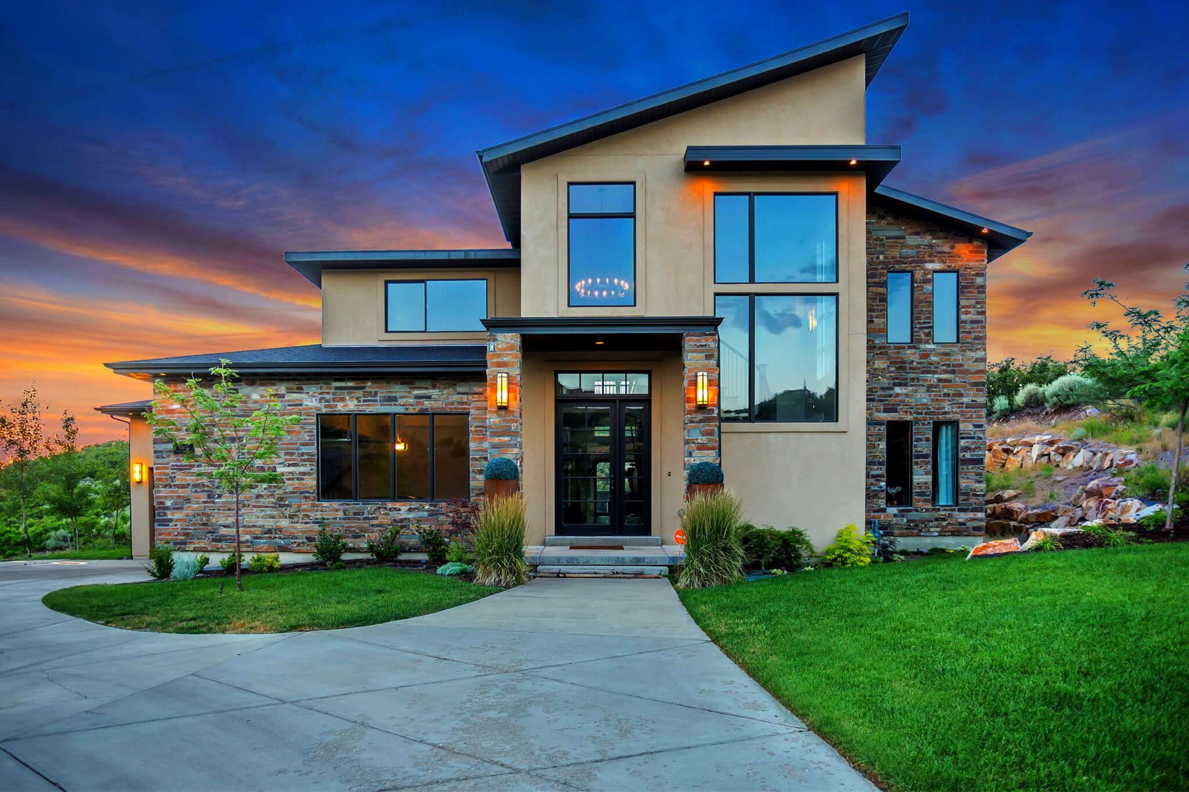 A modern two-story house features a combination of stone and stucco siding, large windows, and a landscaped front yard with a curved driveway.