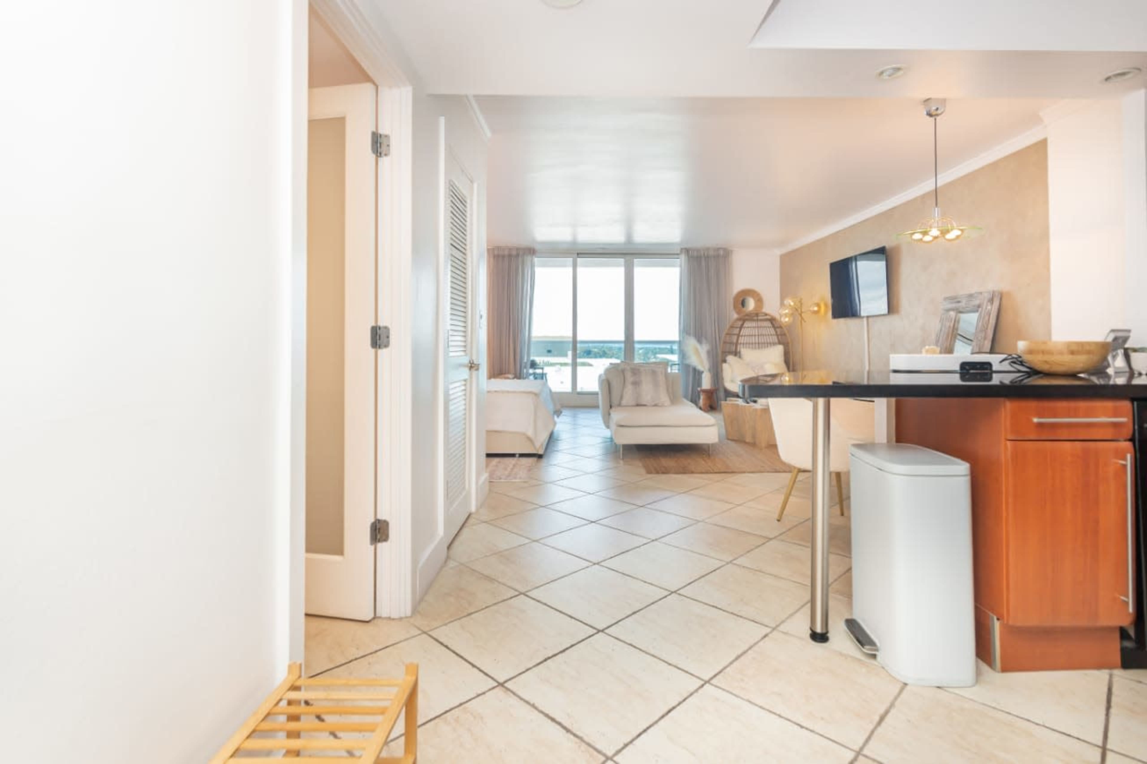 A bright, open room with tiled flooring, featuring a kitchenette, a dining area, and large windows overlooking a view.