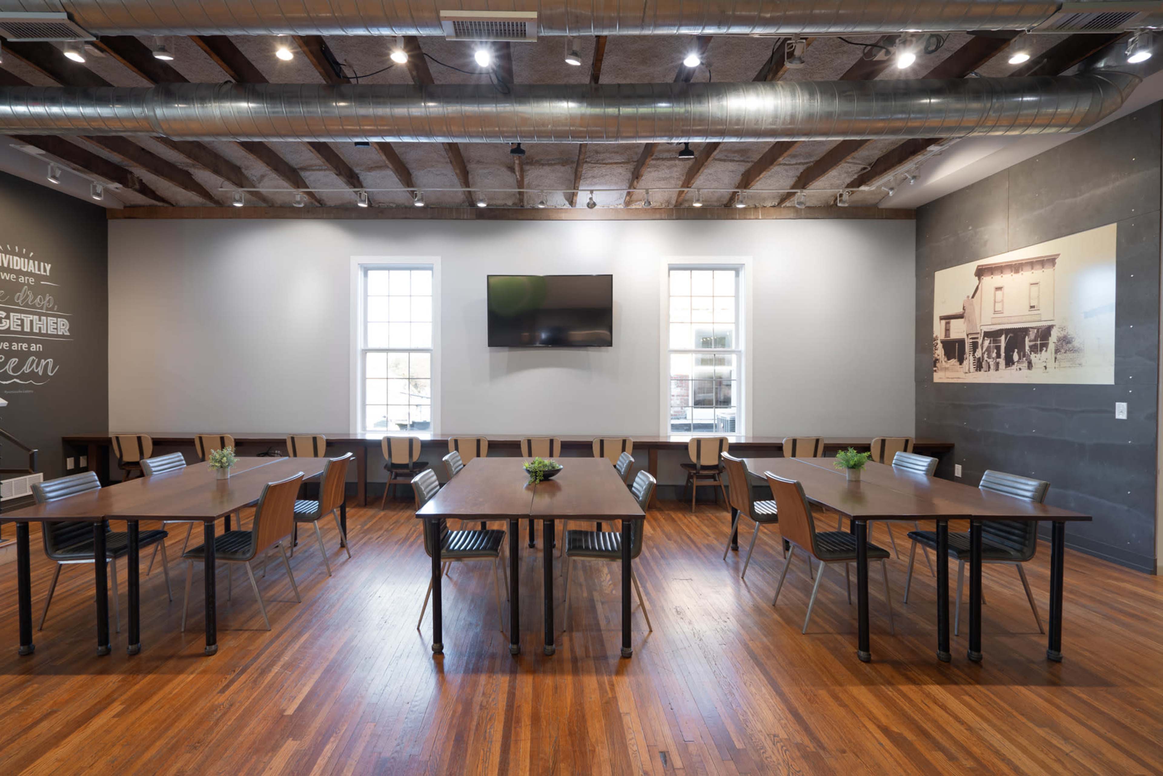 The image shows a spacious dining area with wooden tables and chairs arranged neatly, large windows letting in natural light, and a wall-mounted television.