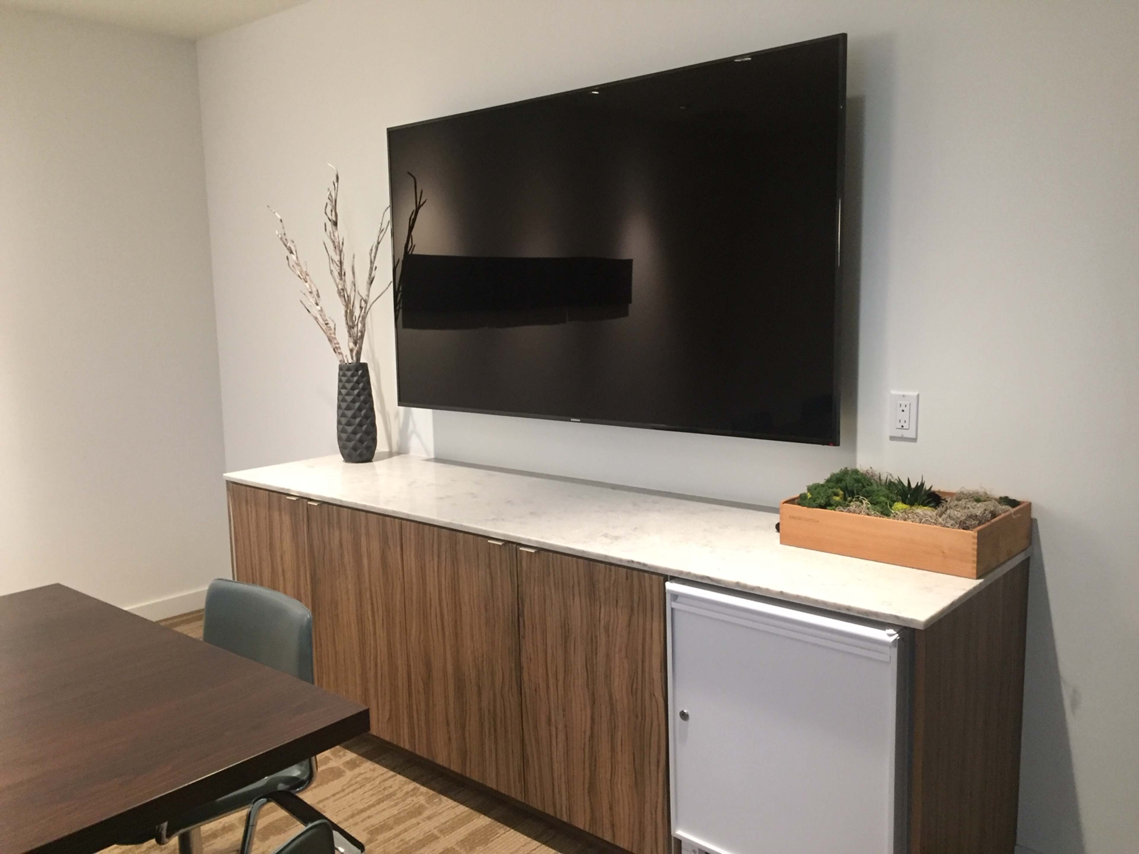 A wall-mounted television is displayed above a wooden cabinet that features a small refrigerator and a planter with greenery.