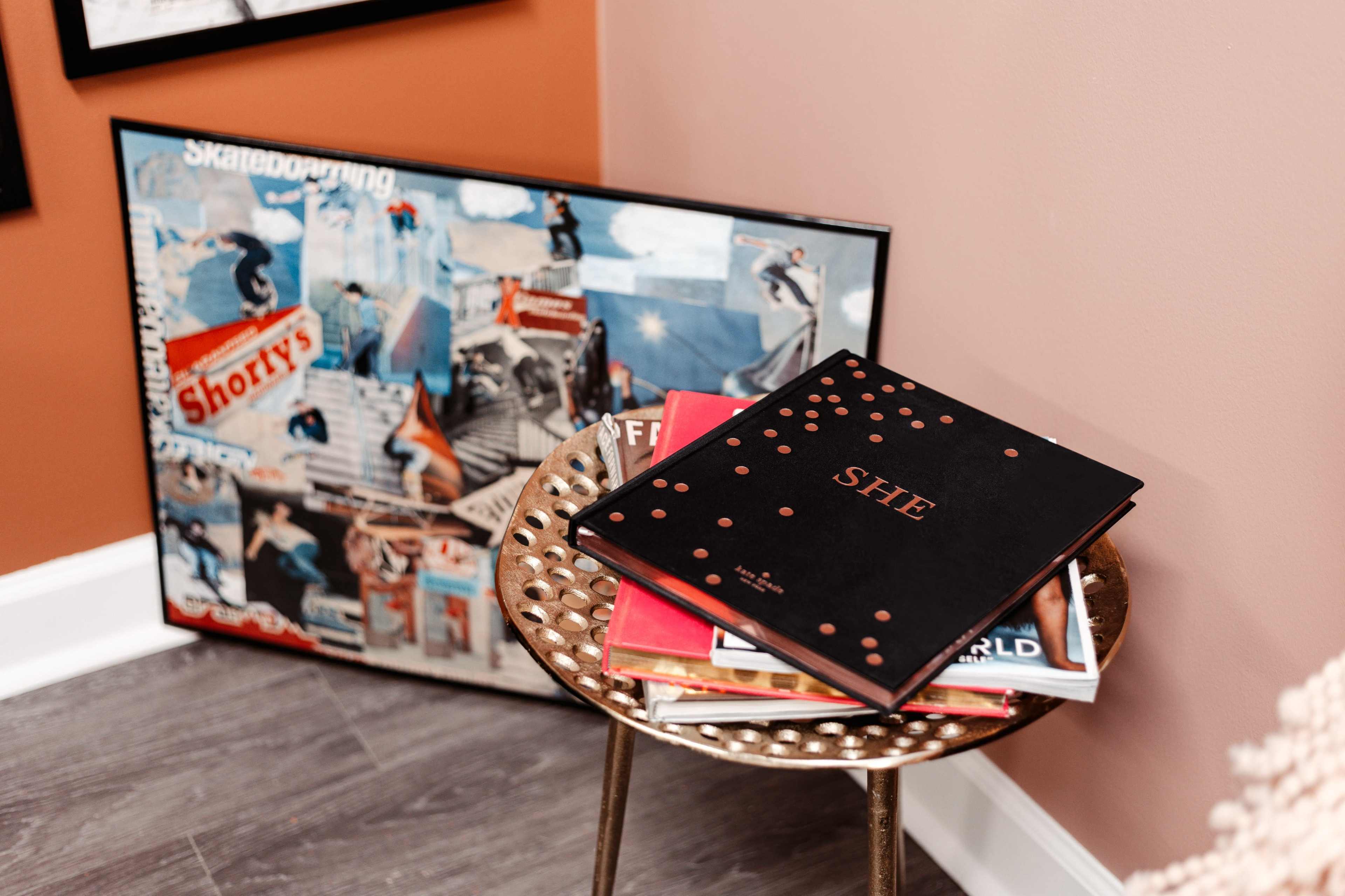 A stack of magazines and a book titled "SHE" rests on a decorative metal side table beside a framed collage of skateboarding images.