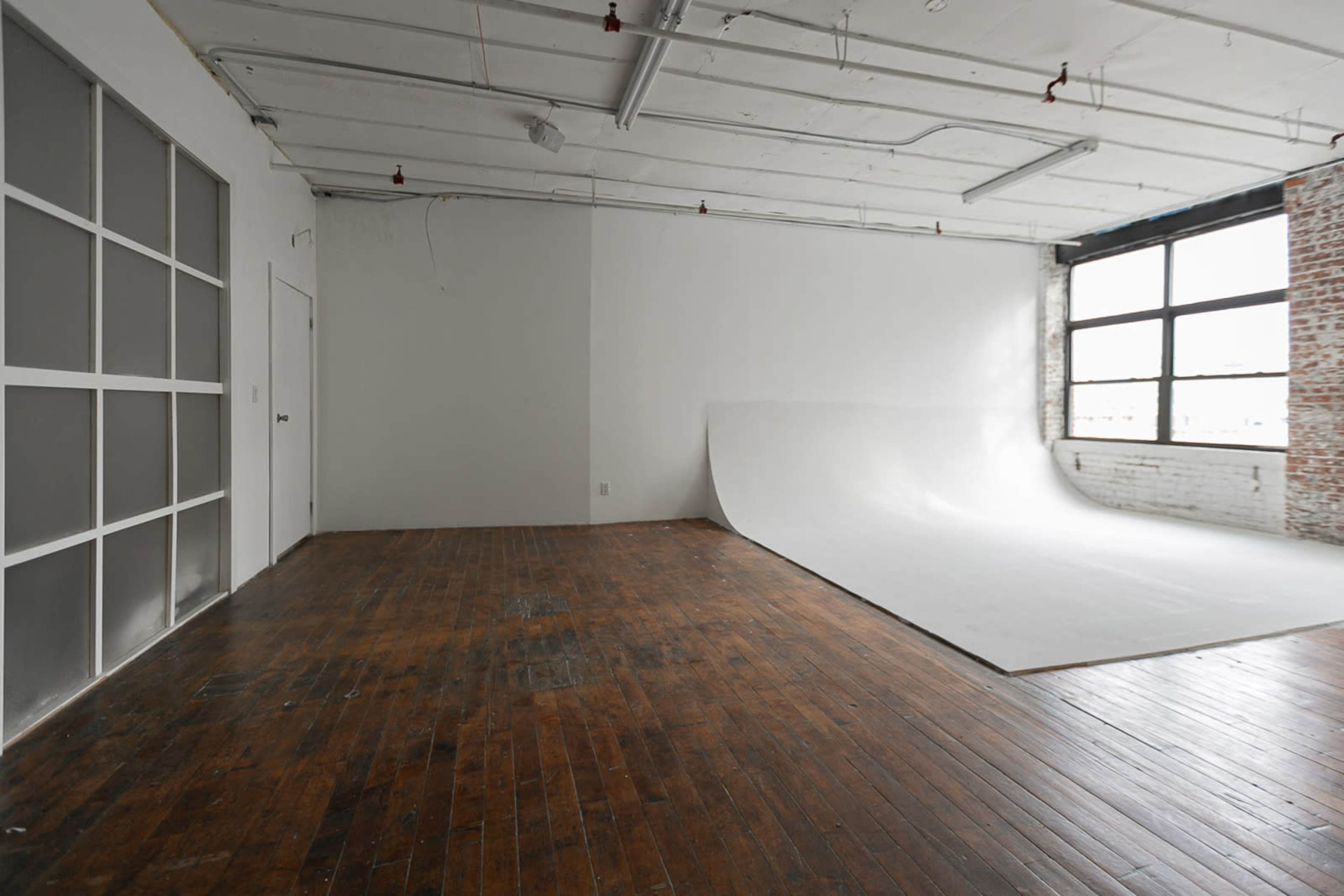 An empty studio space with a white seamless backdrop, wooden flooring, and large windows allowing light to enter.