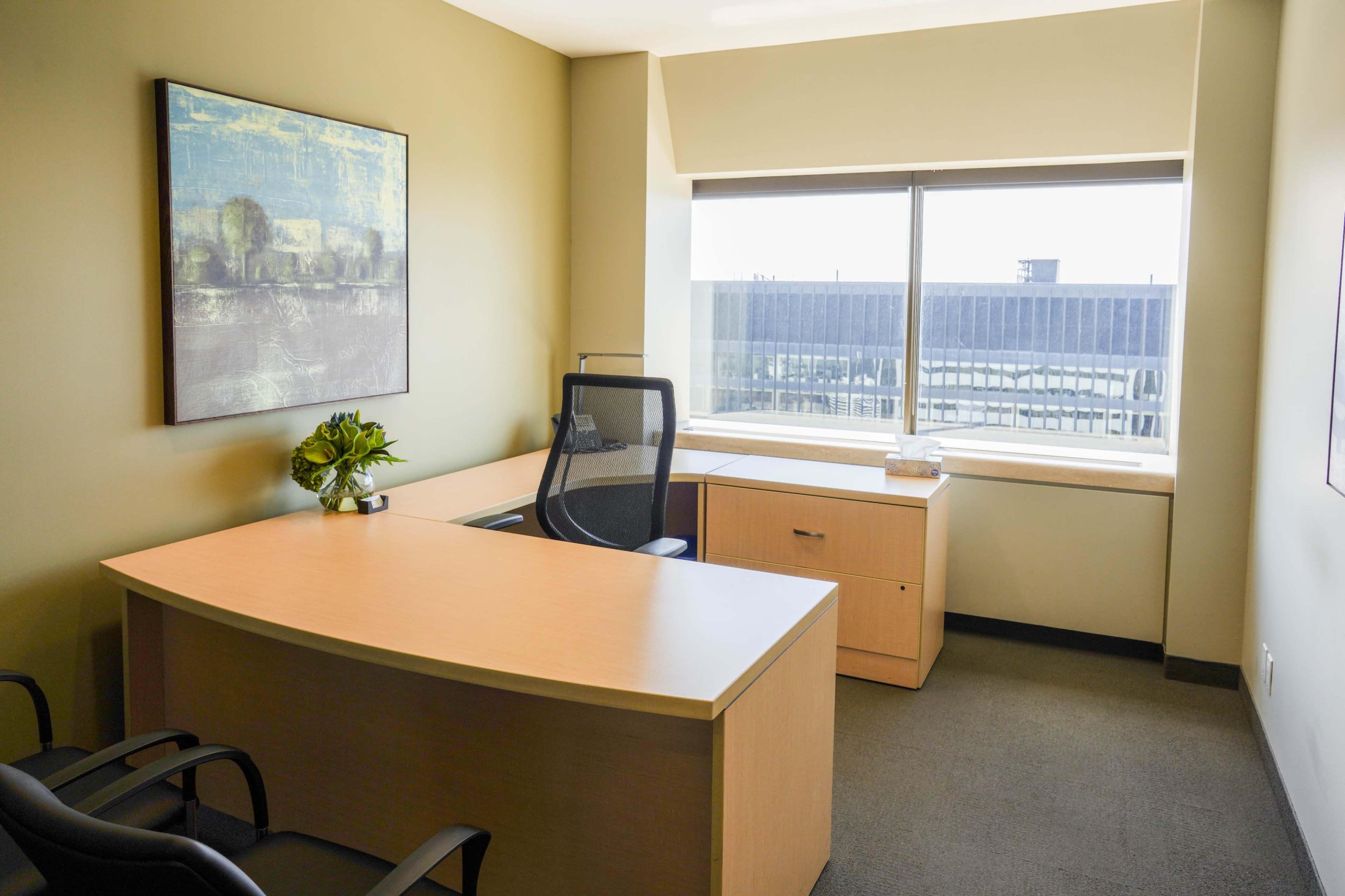 An office space features a wooden desk, a black ergonomic chair, and a large window with a view.