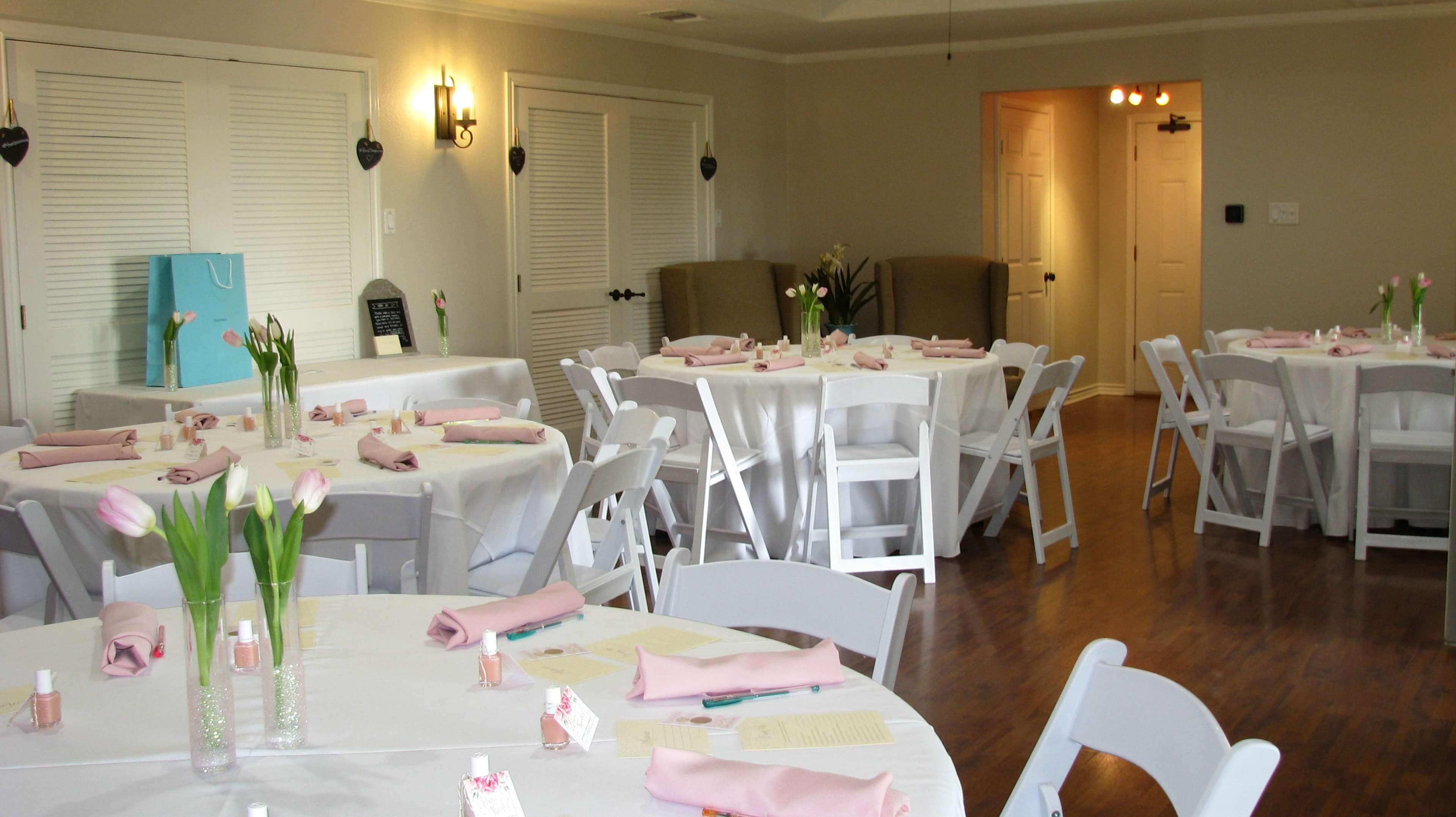 A well-lit event space set up for a gathering, featuring white tablecloths on round tables with pink napkins, vases of tulips, and a few chairs arranged around the space.