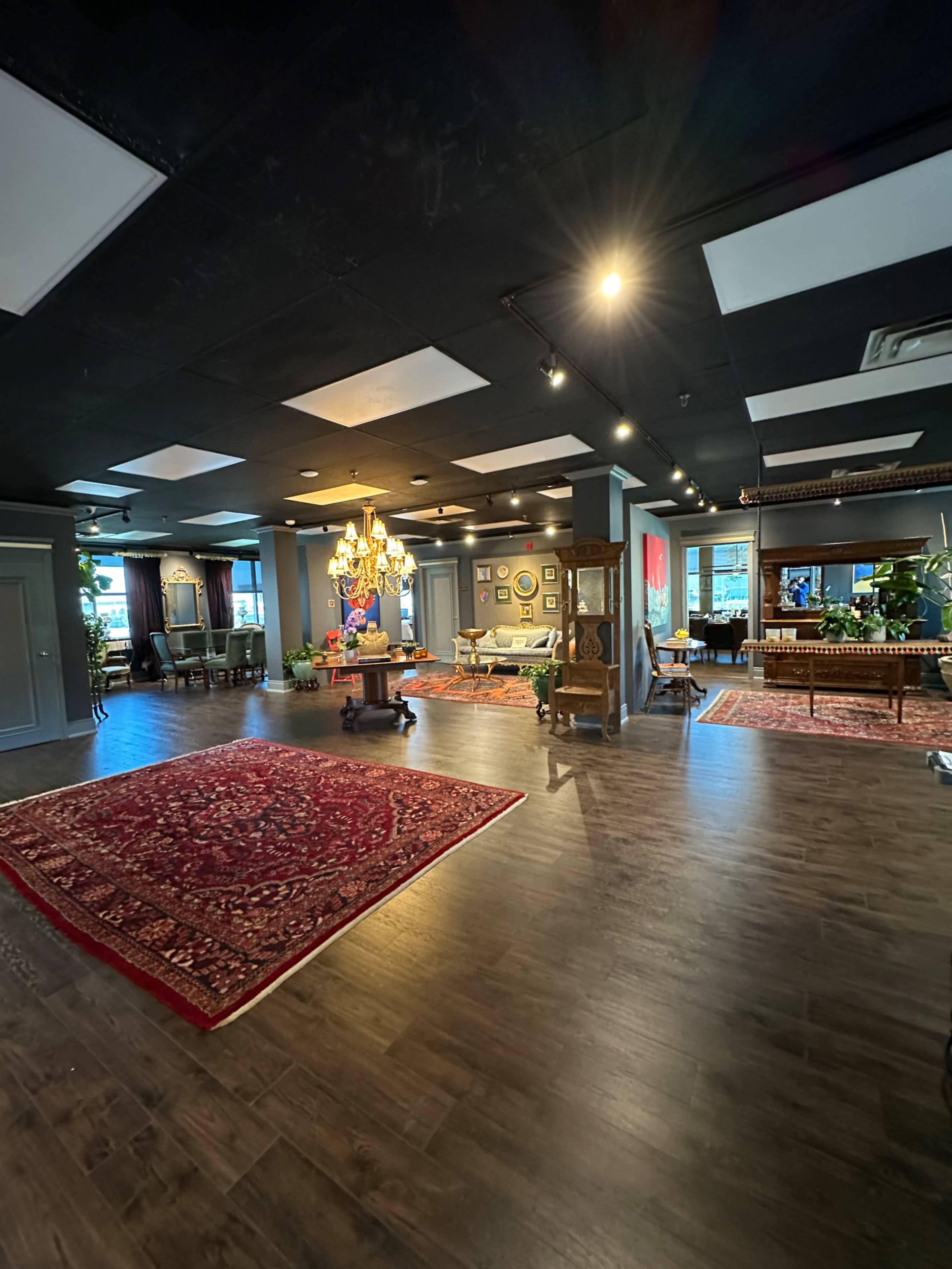 An interior space featuring a large area rug, a chandelier, framed artwork on the walls, and various seating arrangements.