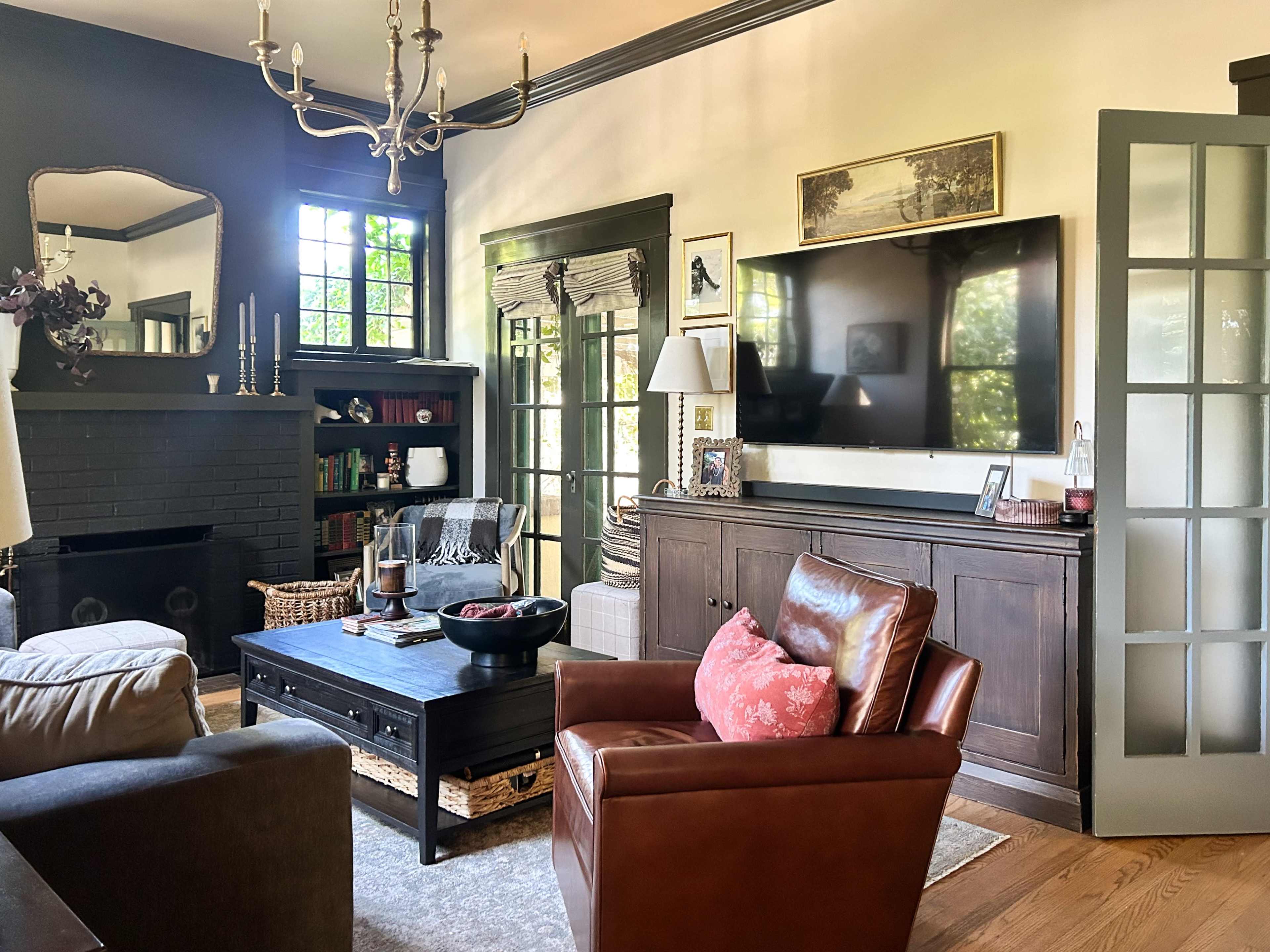 The living room features a mix of vintage and modern furniture, including a brown leather armchair, a large television mounted on the wall, and shelves filled with books and decor.