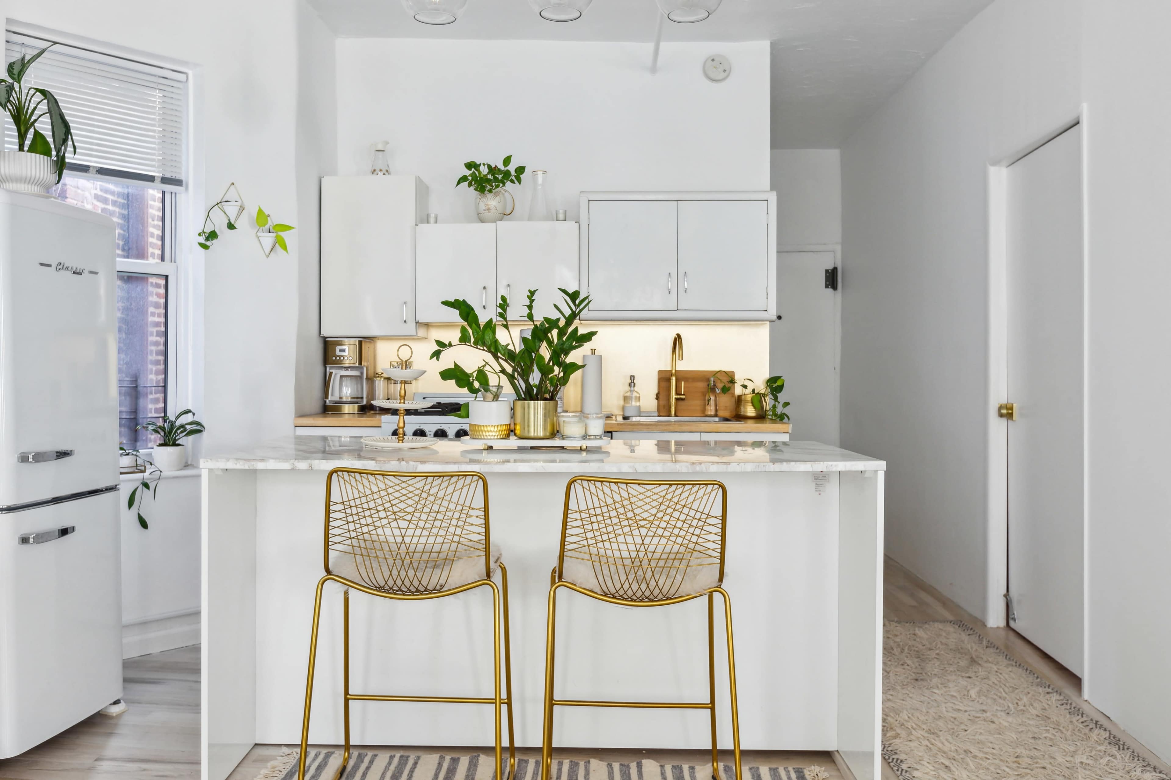 A modern kitchen features a marble-topped island with two gold wire chairs, surrounded by white cabinetry and potted plants.