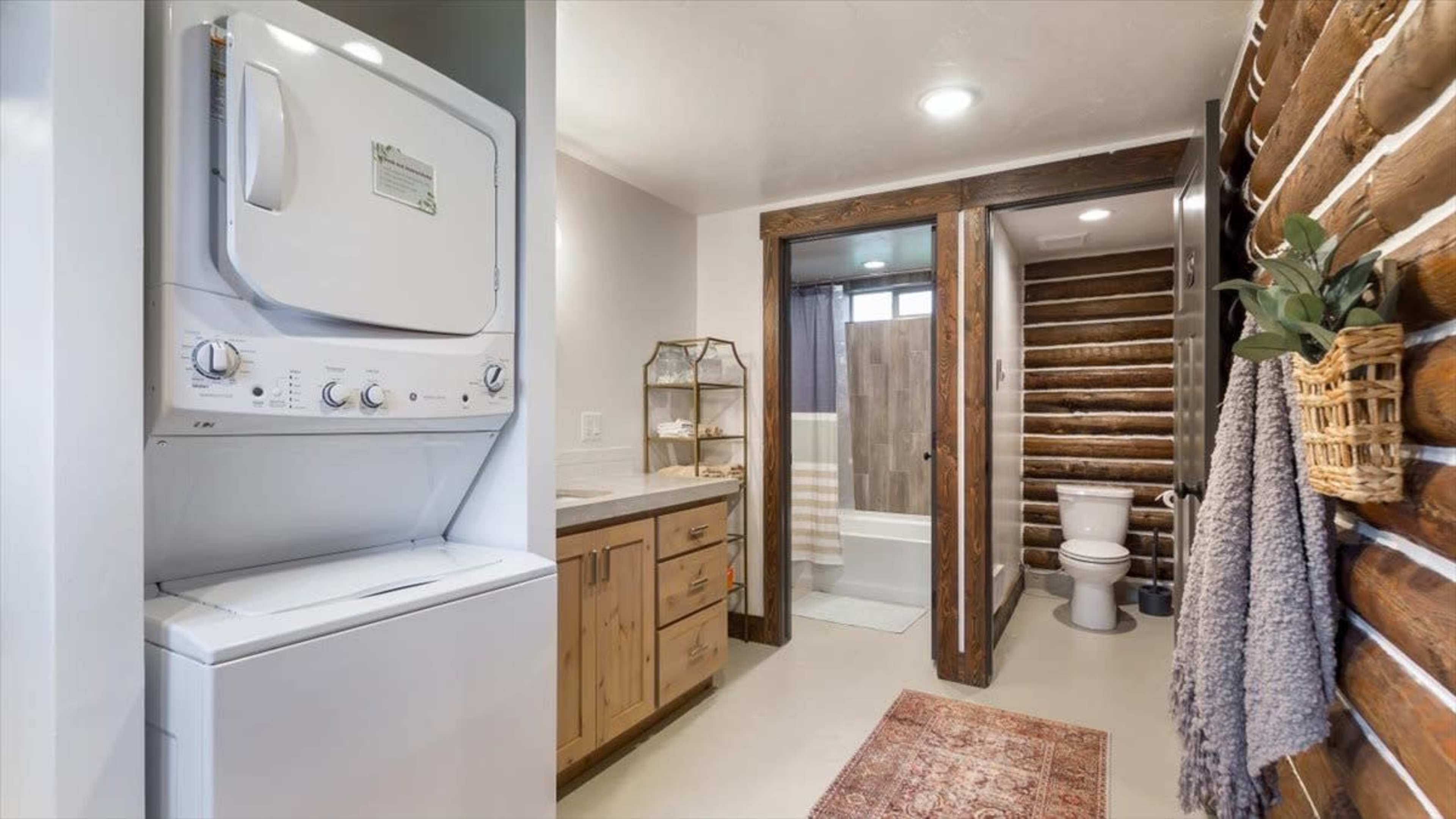 The image shows a laundry area with a stacked washer and dryer, adjacent to a bathroom featuring a shower, wooden accents, and a toilet.
