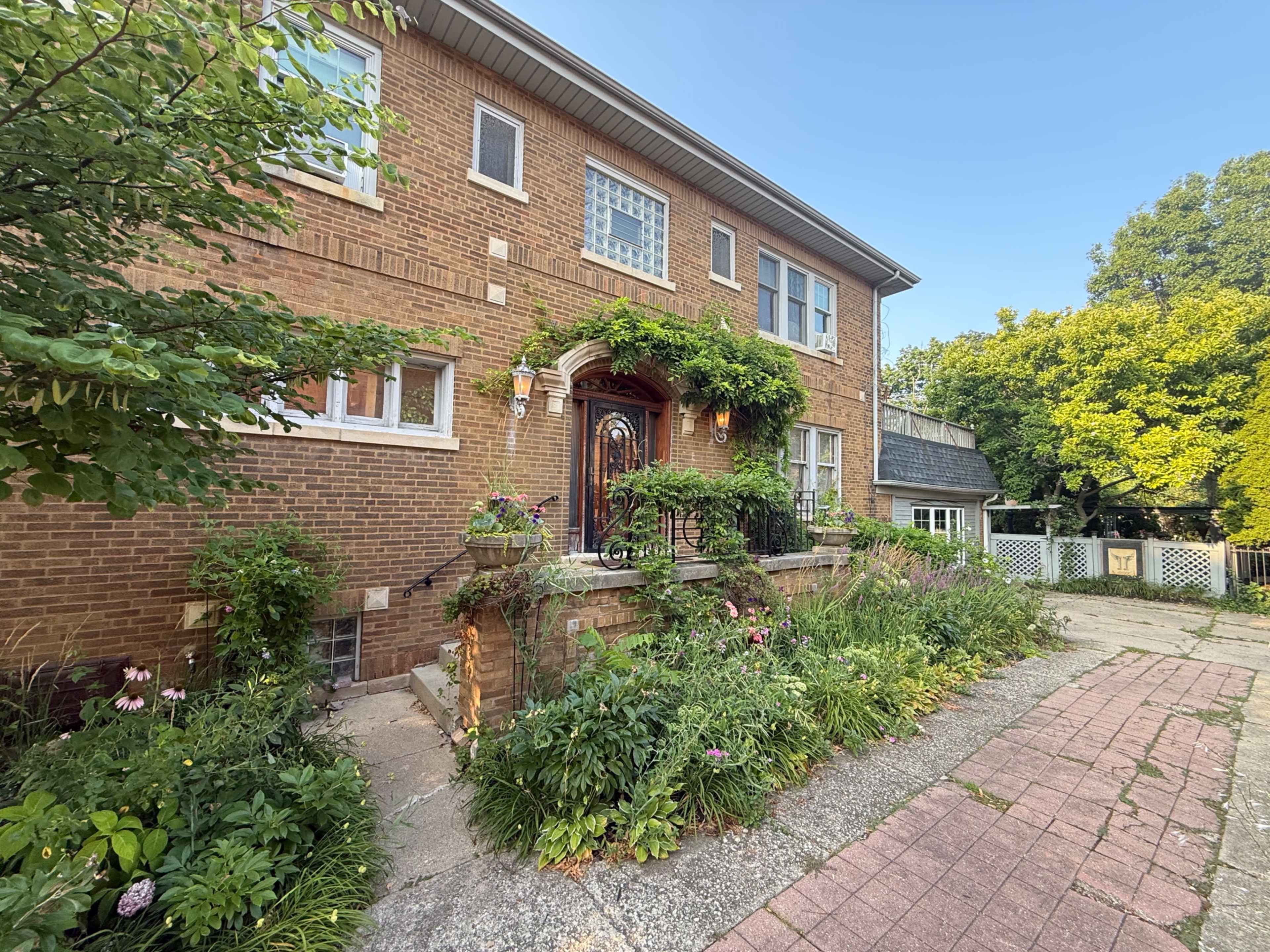 The image shows a two-story brick house with a landscaped front garden featuring various plants and flowers.