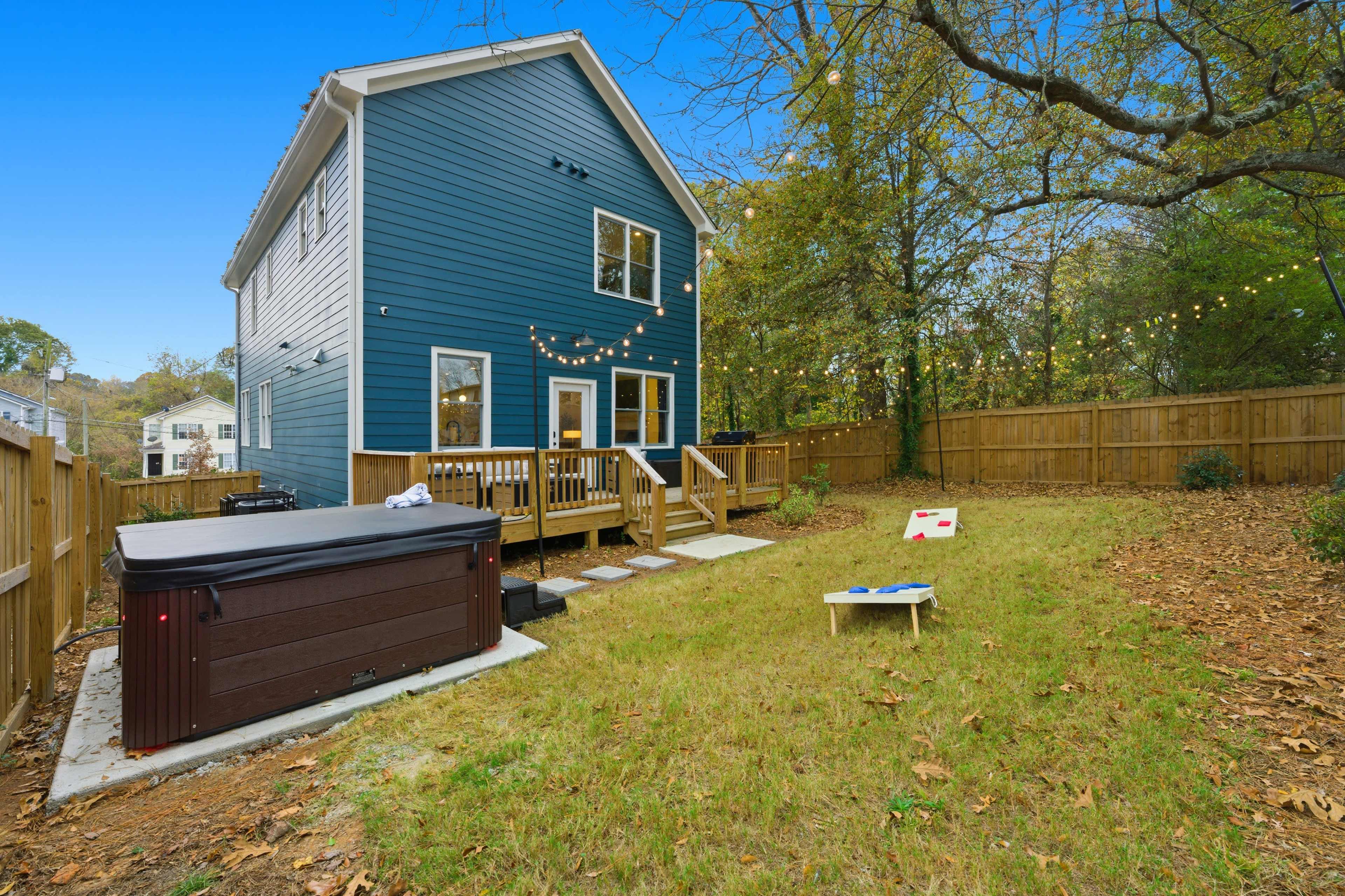 A two-story blue house with a fenced backyard features a hot tub, a wooden deck, and a cornhole game set on the lawn.
