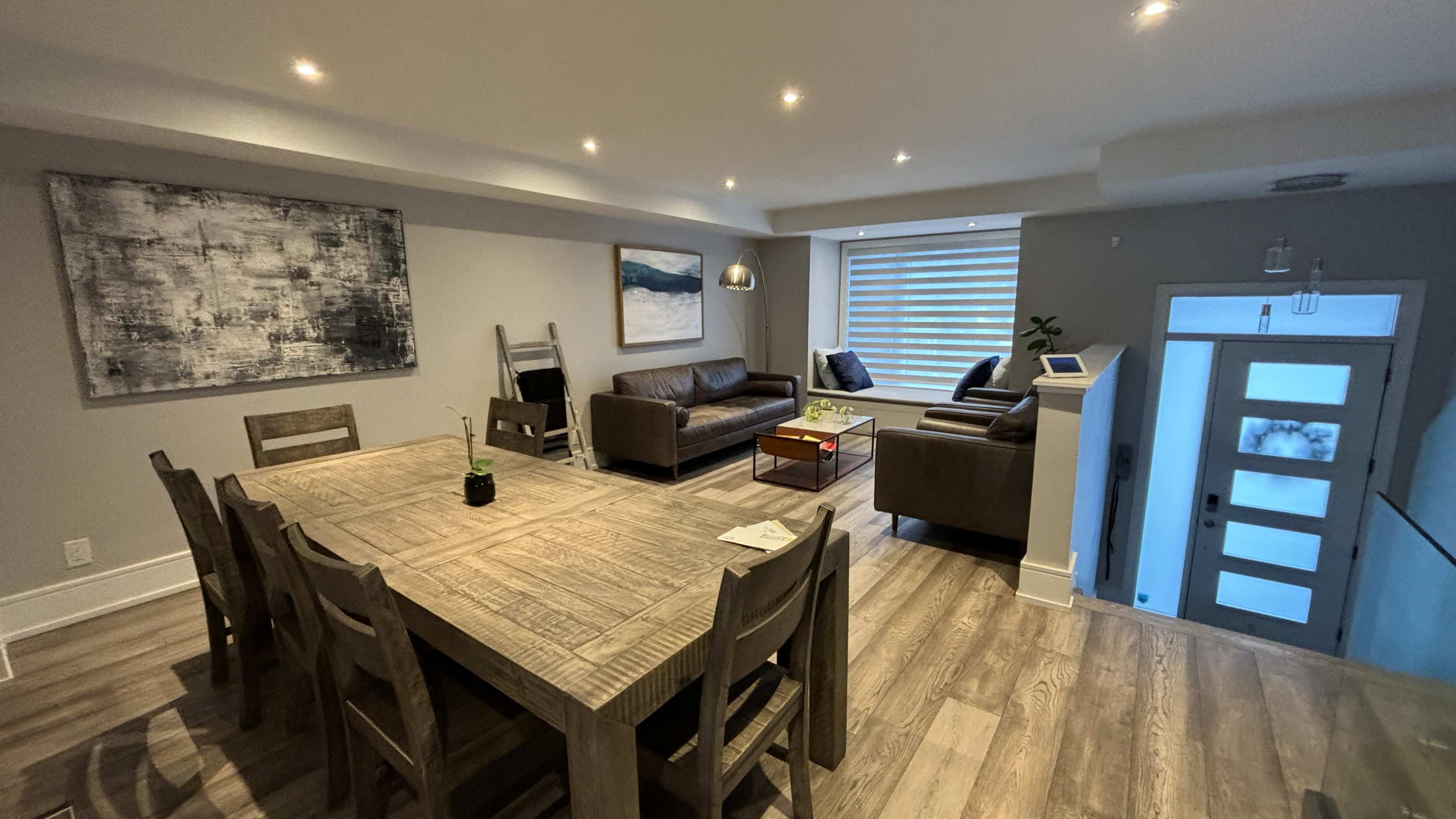 A modern living room and dining area with wooden furniture, two brown sofas, and a large dining table, all set in a light-colored space.