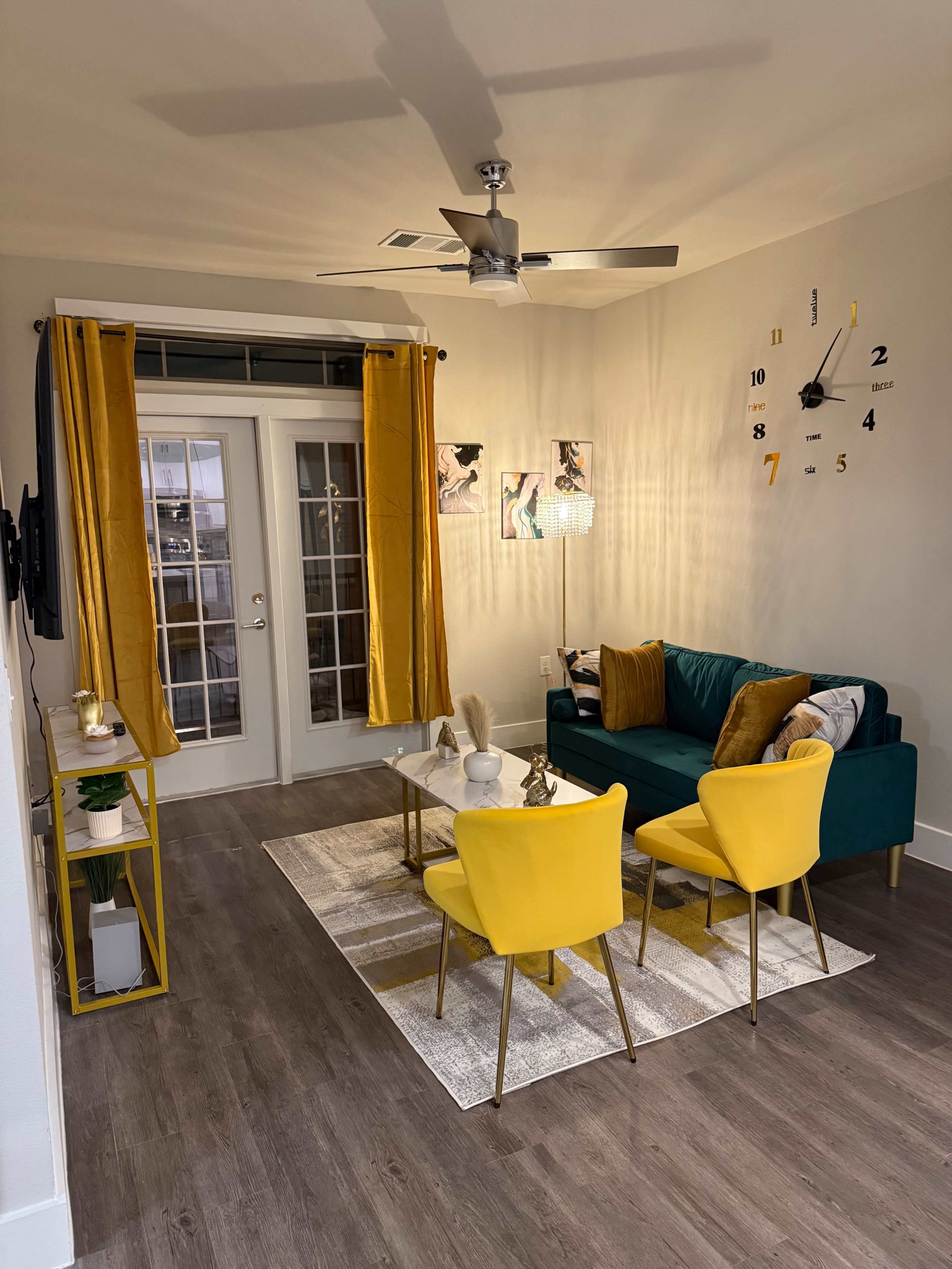The image shows a modern living room featuring a teal sofa, two yellow chairs, a coffee table, and large windows with sheer curtains.