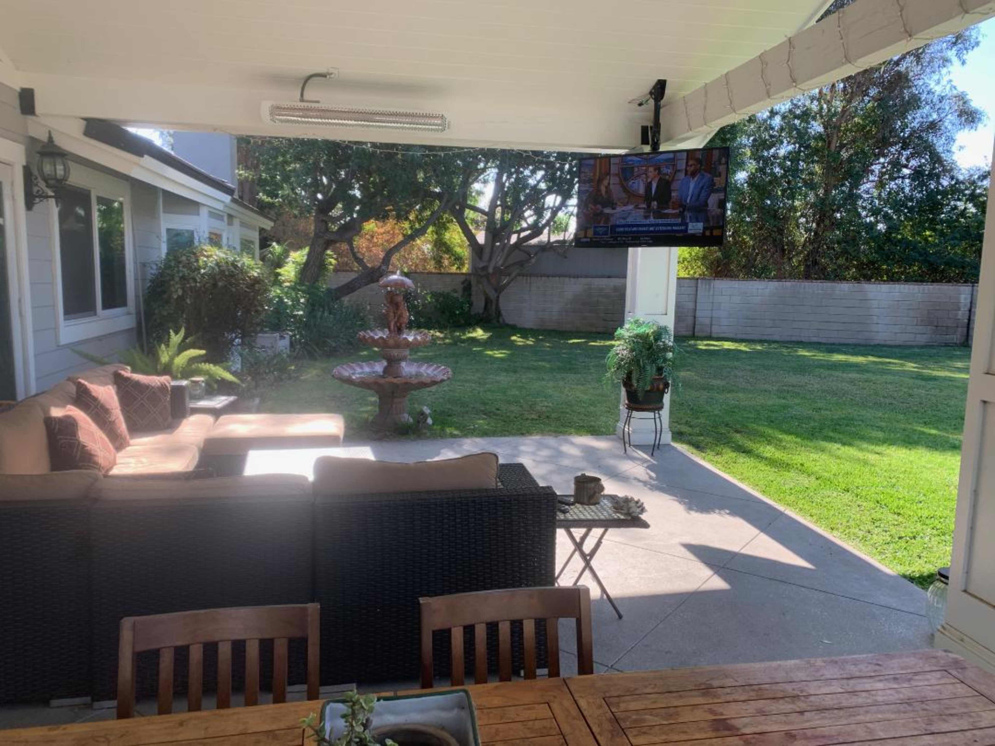 A covered outdoor patio area with a seating arrangement, a decorative fountain, a television mounted on the wall, and a spacious lawn beyond.