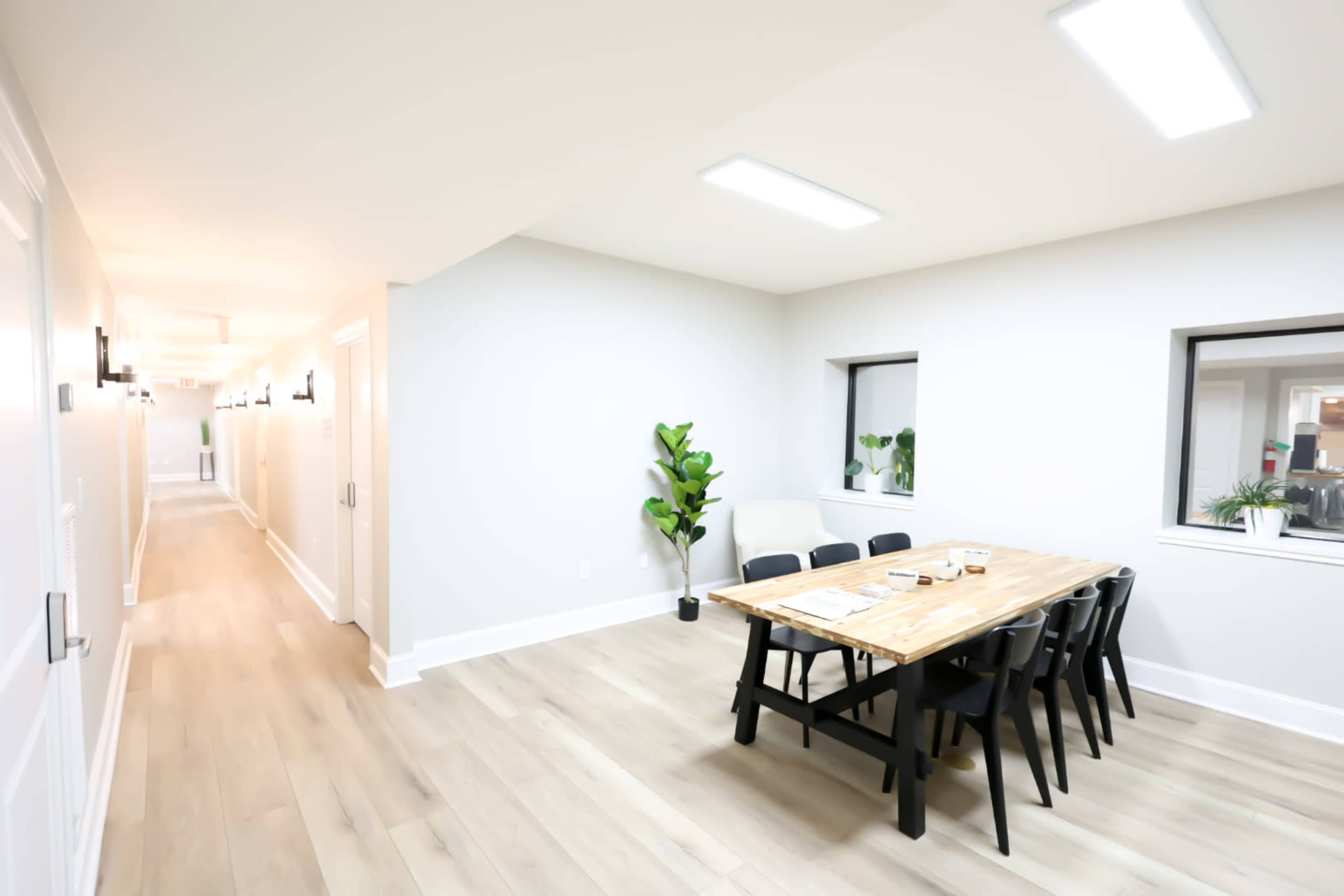 A hallway leads to a brightly lit room with a wooden dining table and black chairs, accompanied by a small plant and two windows.