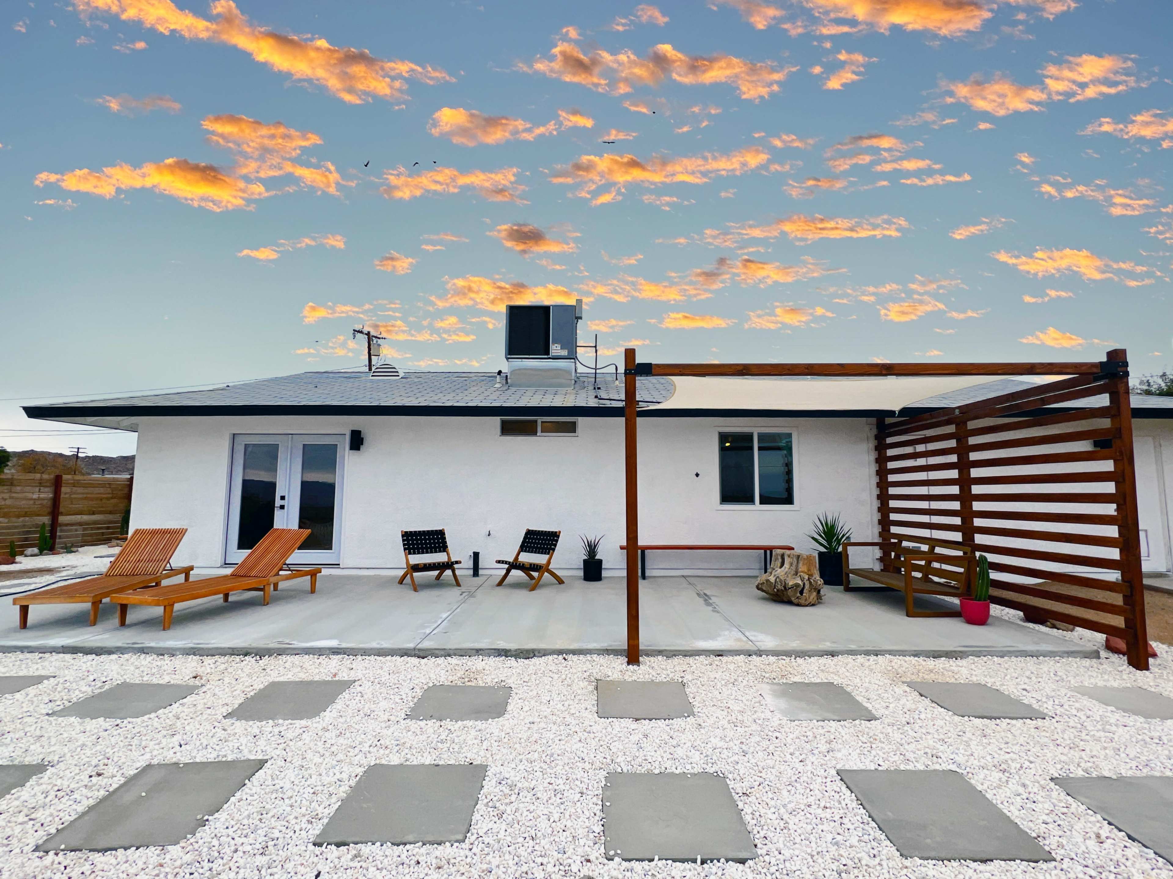 A modern house with a patio featuring two lounge chairs, a wooden pergola, and a landscaped area of gravel and stepping stones under a colorful sky.