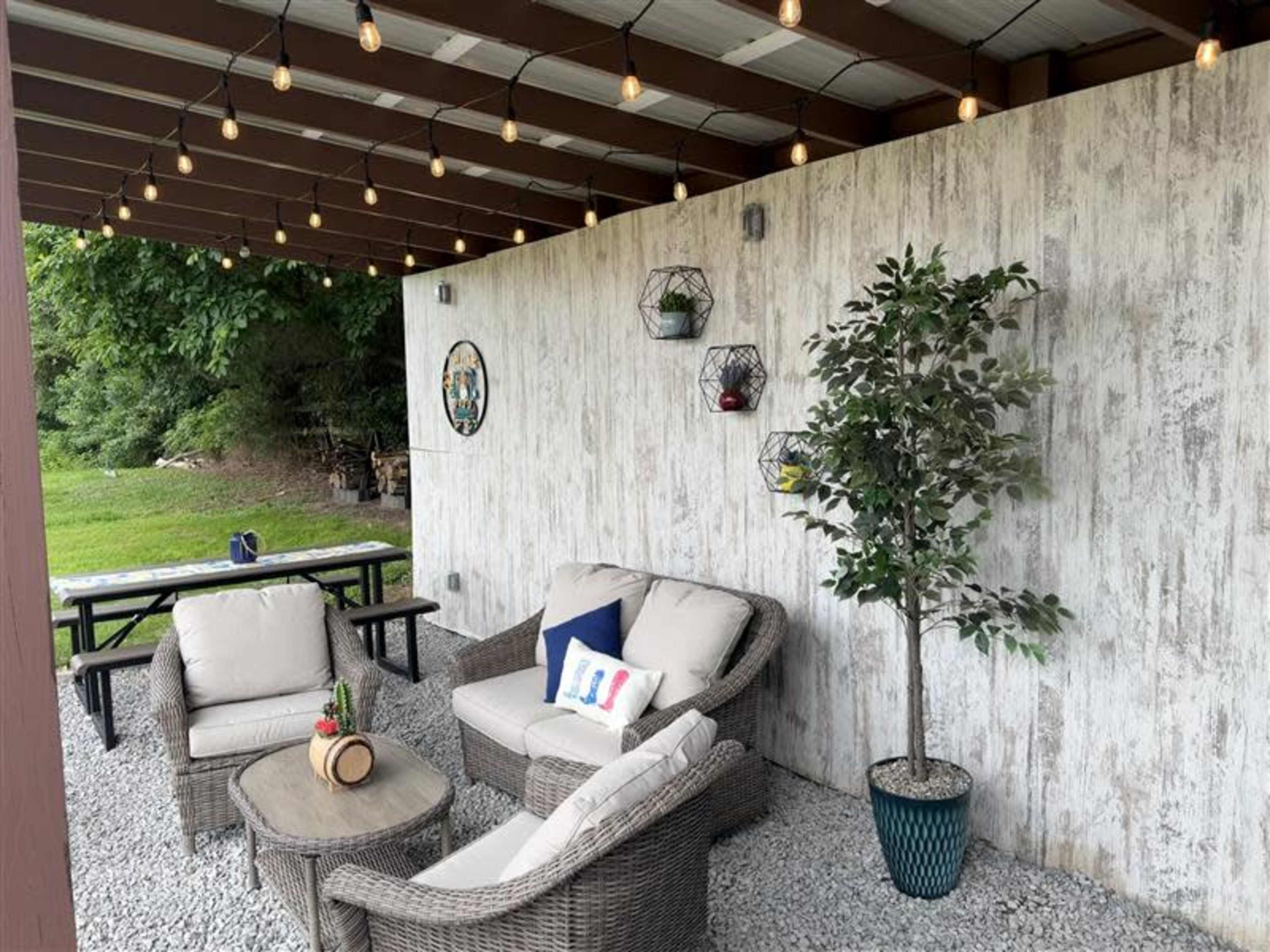 A cozy outdoor seating area features two plush chairs, a small table, and a decorative plant under string lights and backed by a rustic wall.