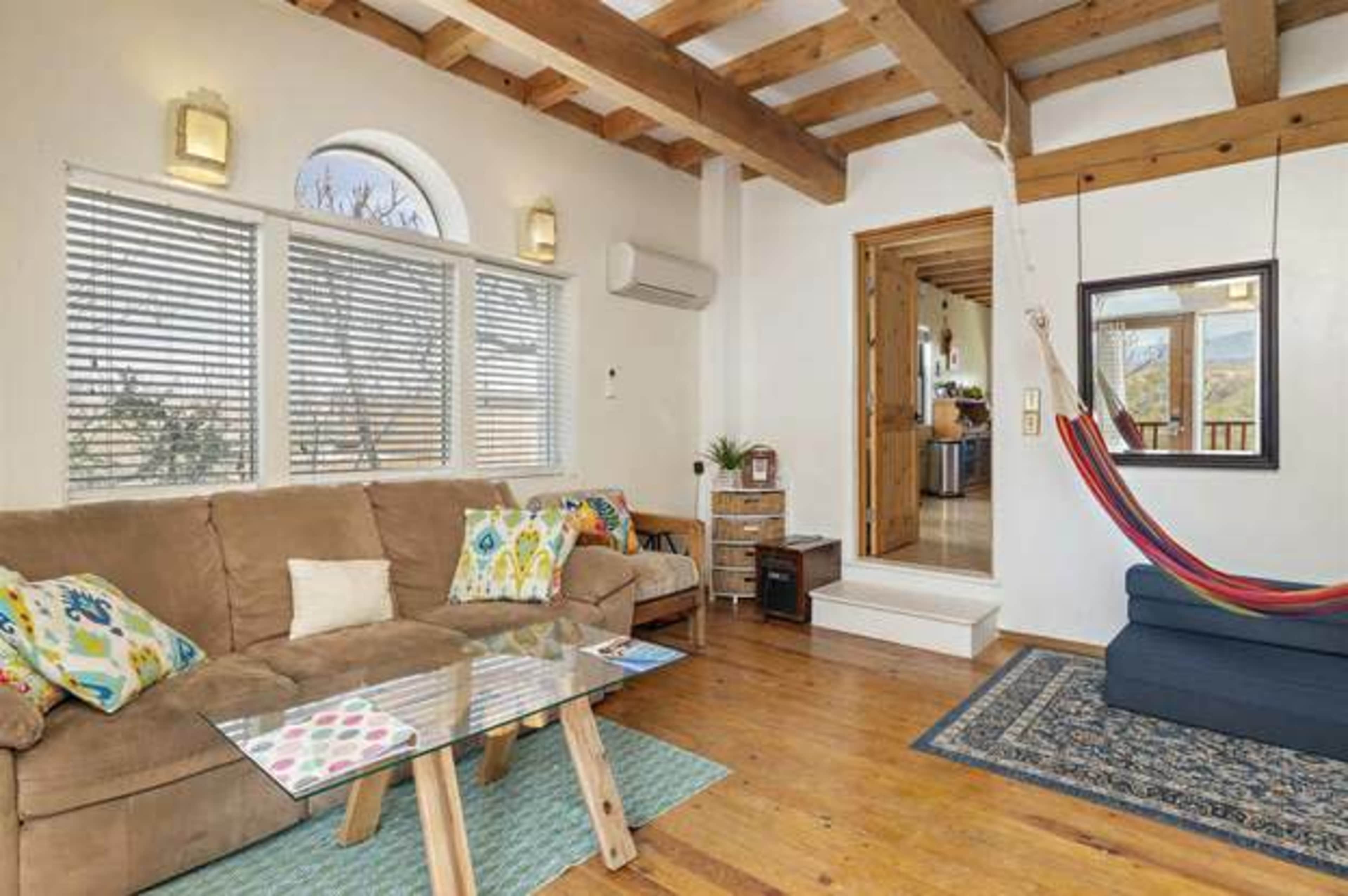 A cozy living room with a sofa, a small glass table, and a hammock, featuring wooden beams in the ceiling and a view through large windows.