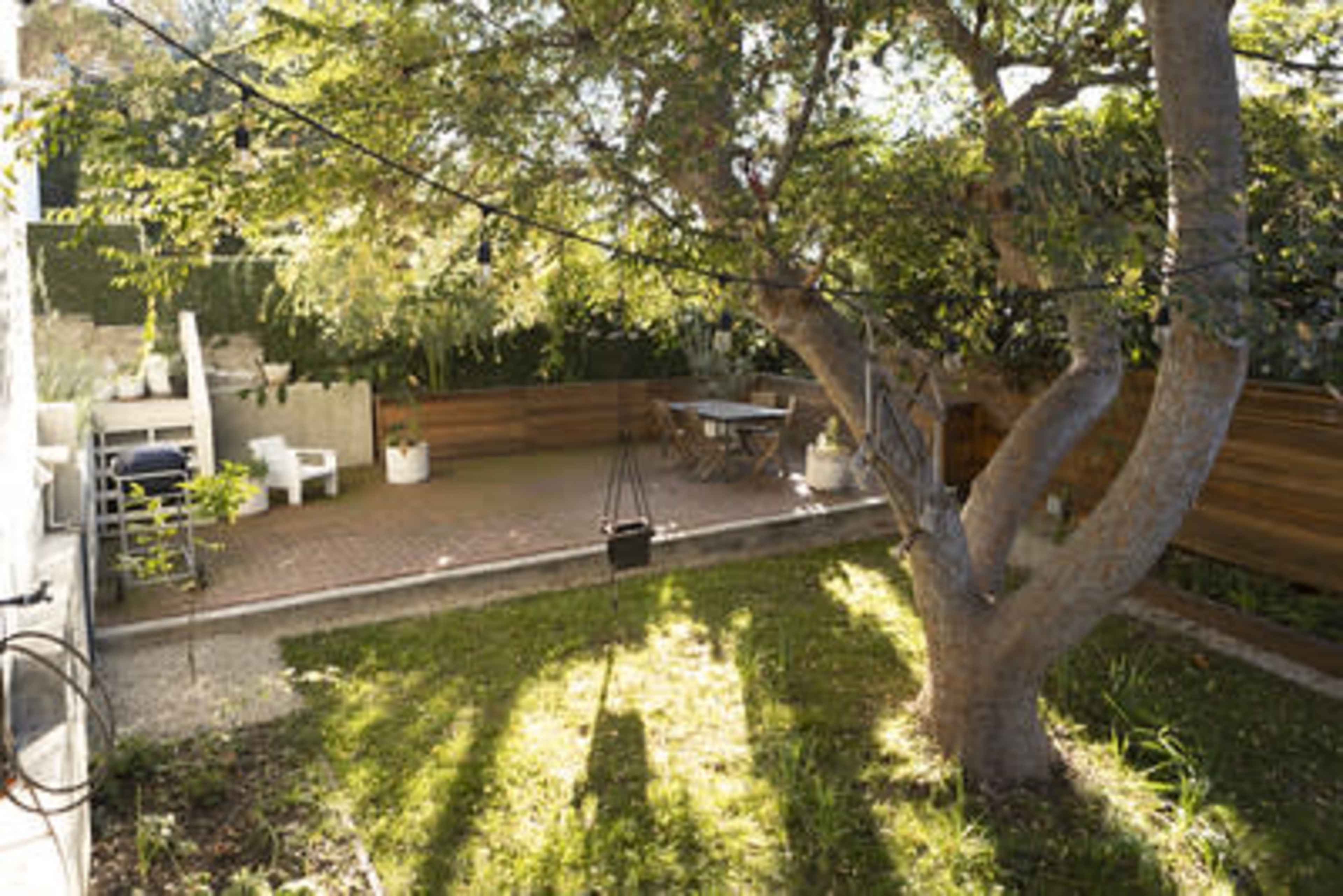 A backyard features a large tree, a brick patio with a table and chairs, and various planters.