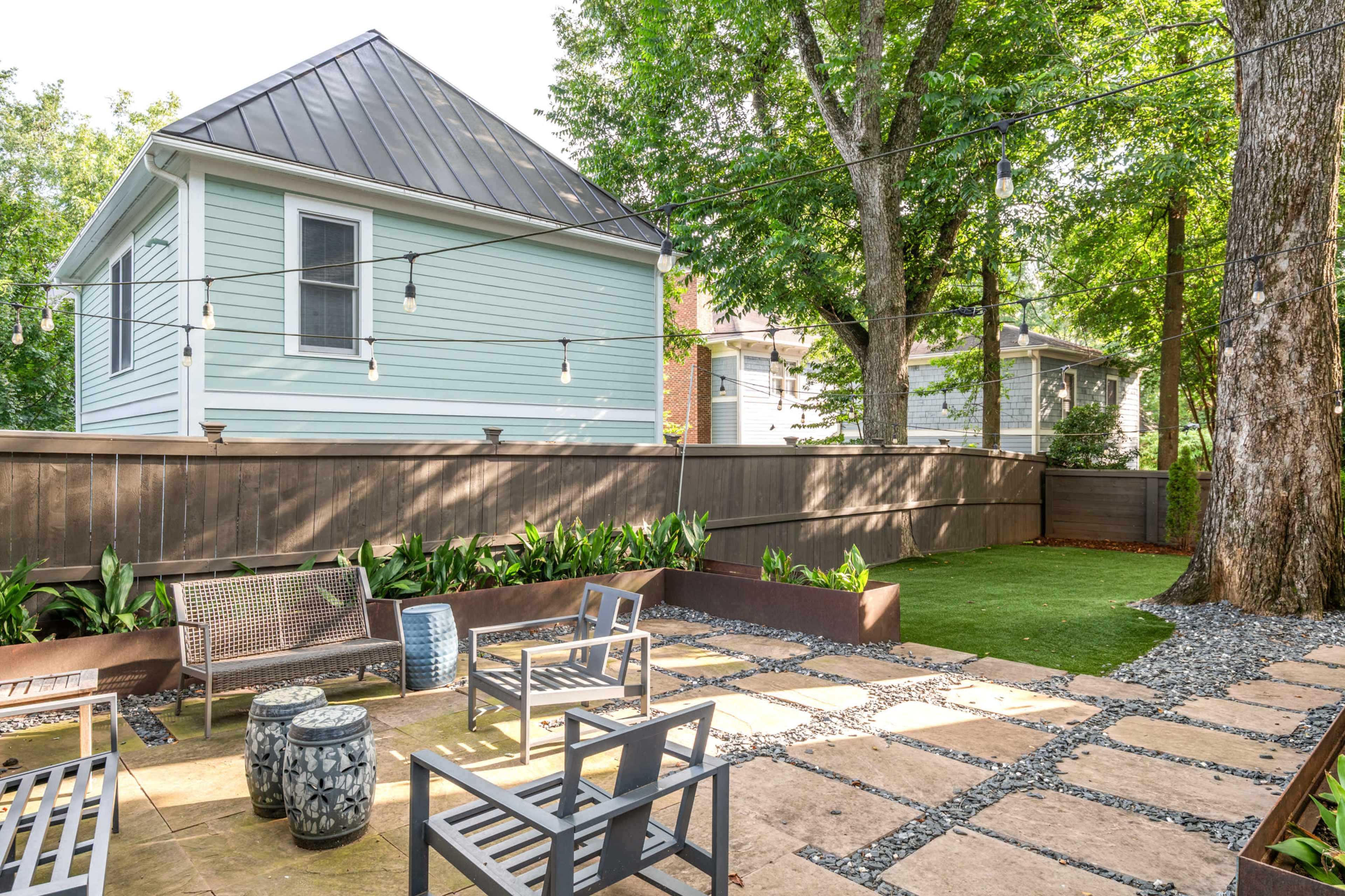A small backyard features a stone patio with seating, bordered by a wooden fence and greenery, under overhead string lights.