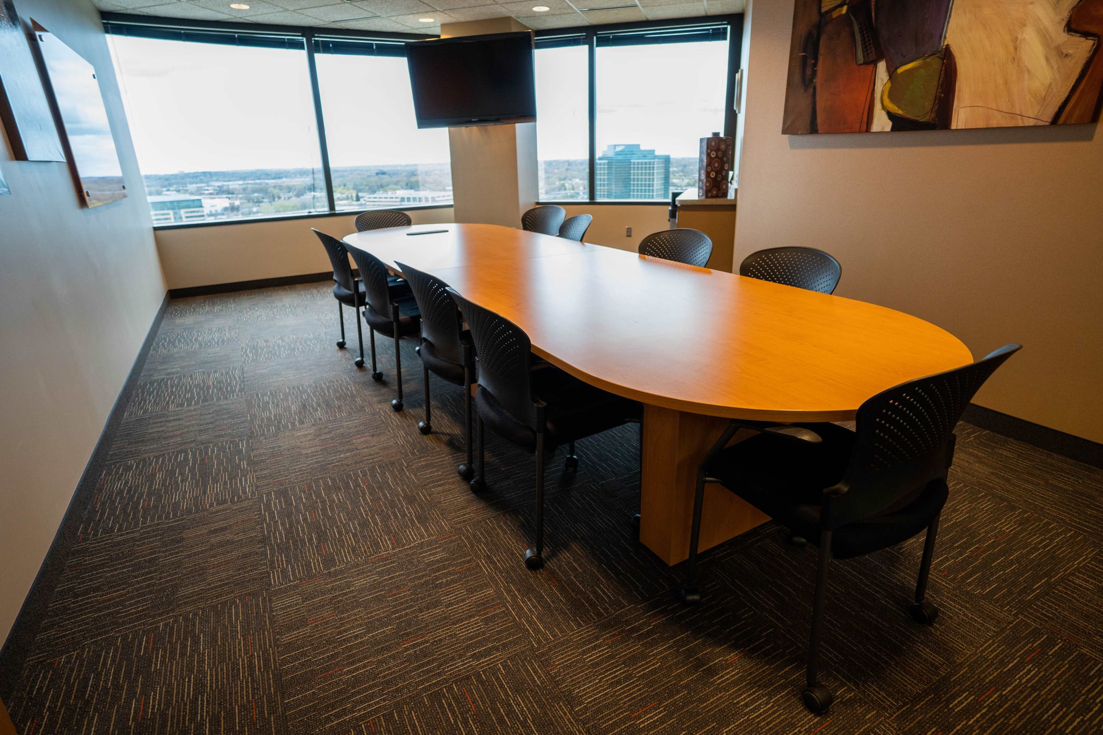 A long, oval conference table with black chairs is positioned in a modern meeting room surrounded by large windows.