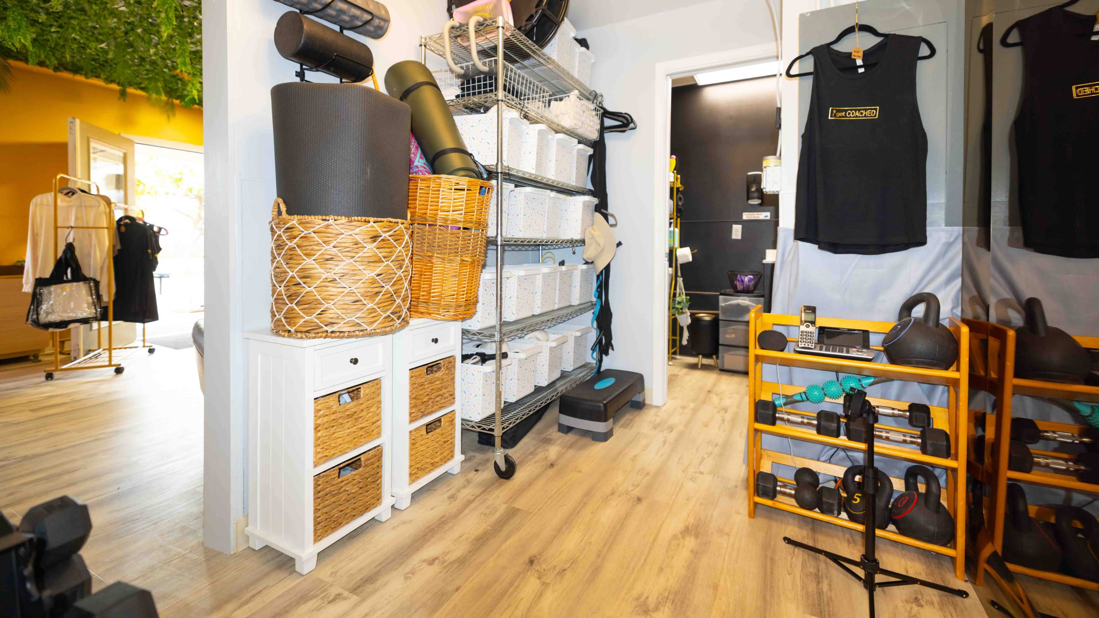 The image shows a fitness studio interior featuring shelves with exercise equipment, storage baskets, and neatly organized workout mats.
