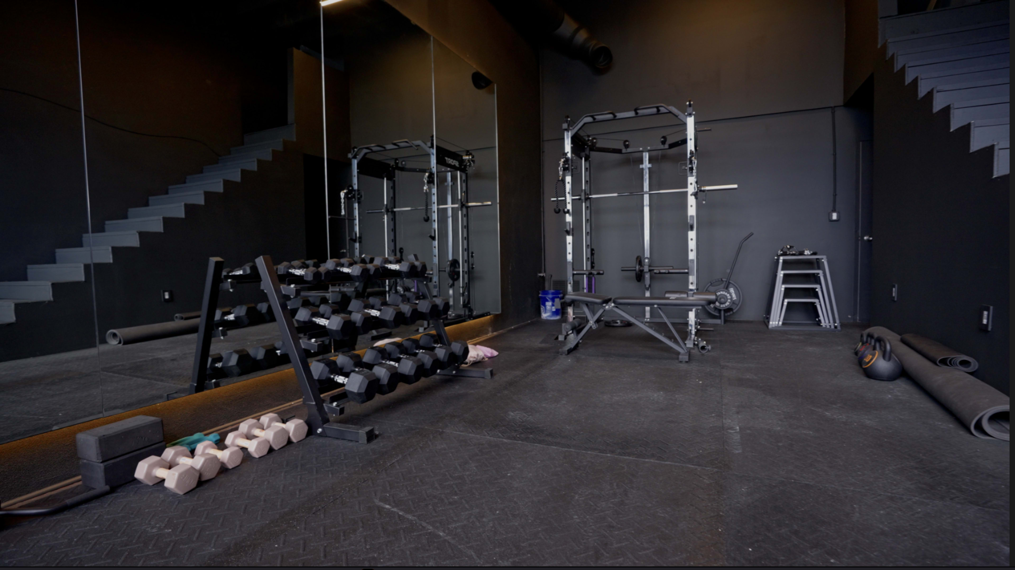 The image shows a well-equipped gym with a squat rack, free weights, and exercise mats, situated in a room with dark walls and large mirrors.