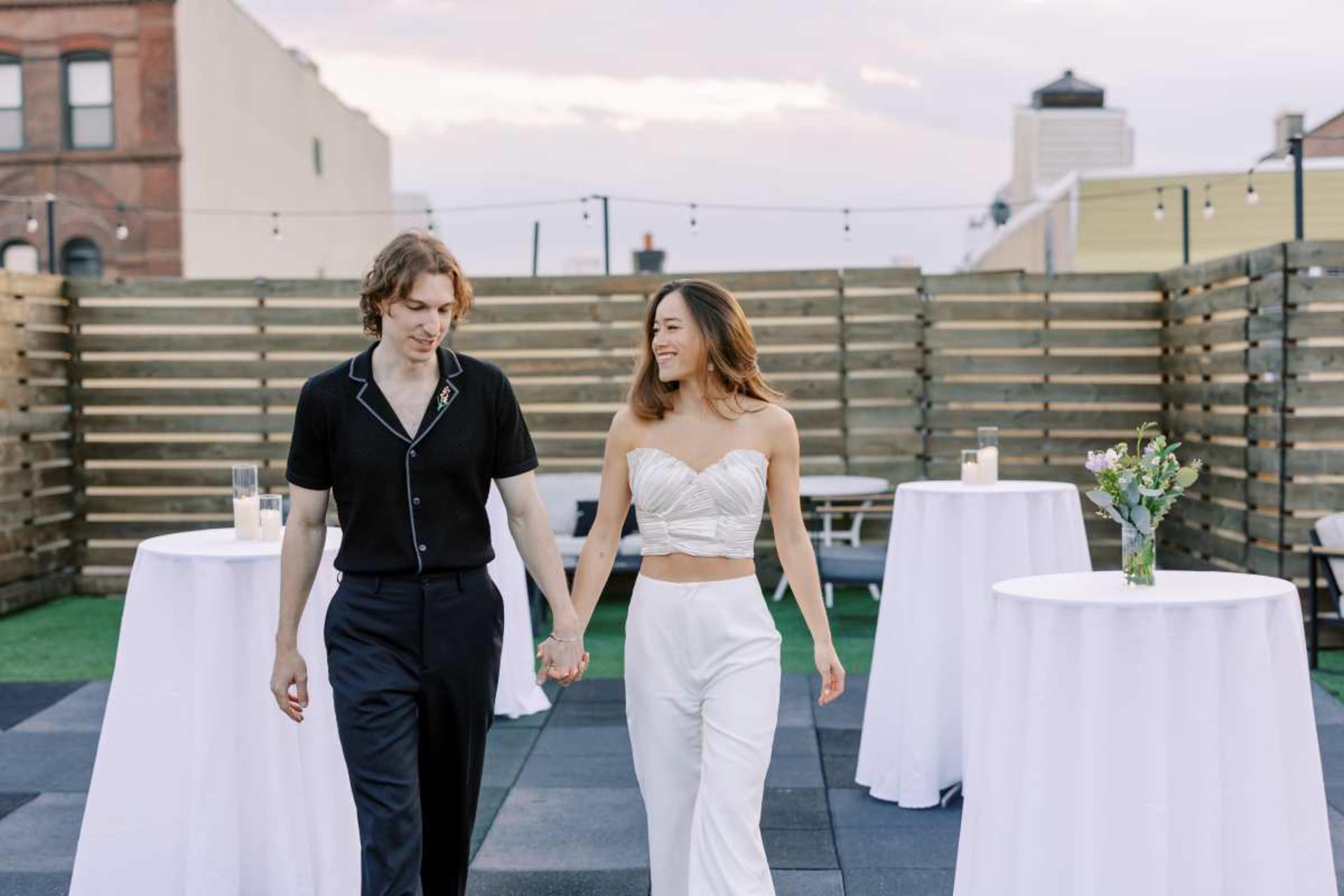 A couple walks hand in hand on a rooftop, surrounded by small tables and decorative greenery under a pastel sky.