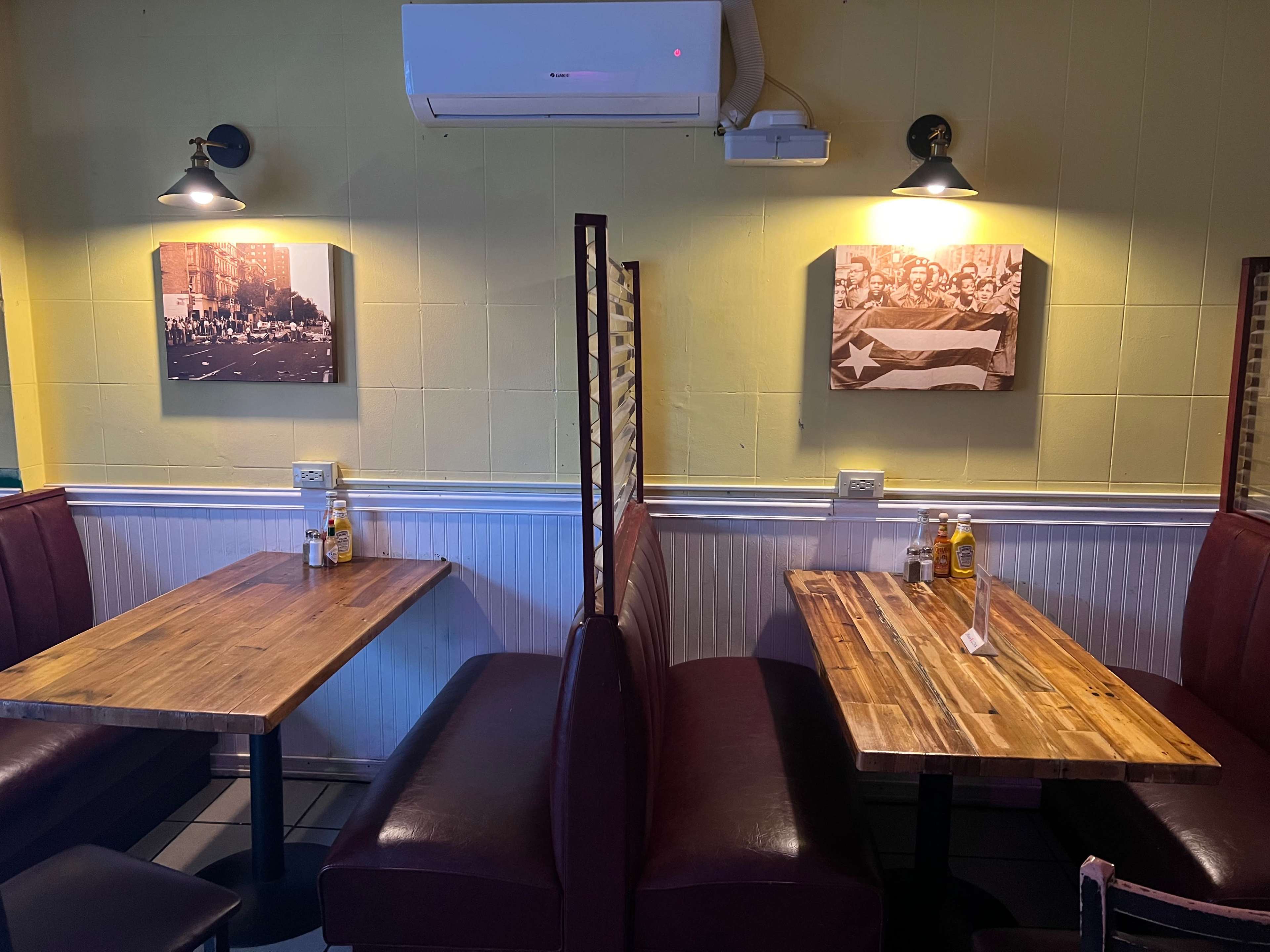 An interior of a restaurant featuring two wooden tables with maroon upholstered seating and framed black-and-white photos on the yellow walls.