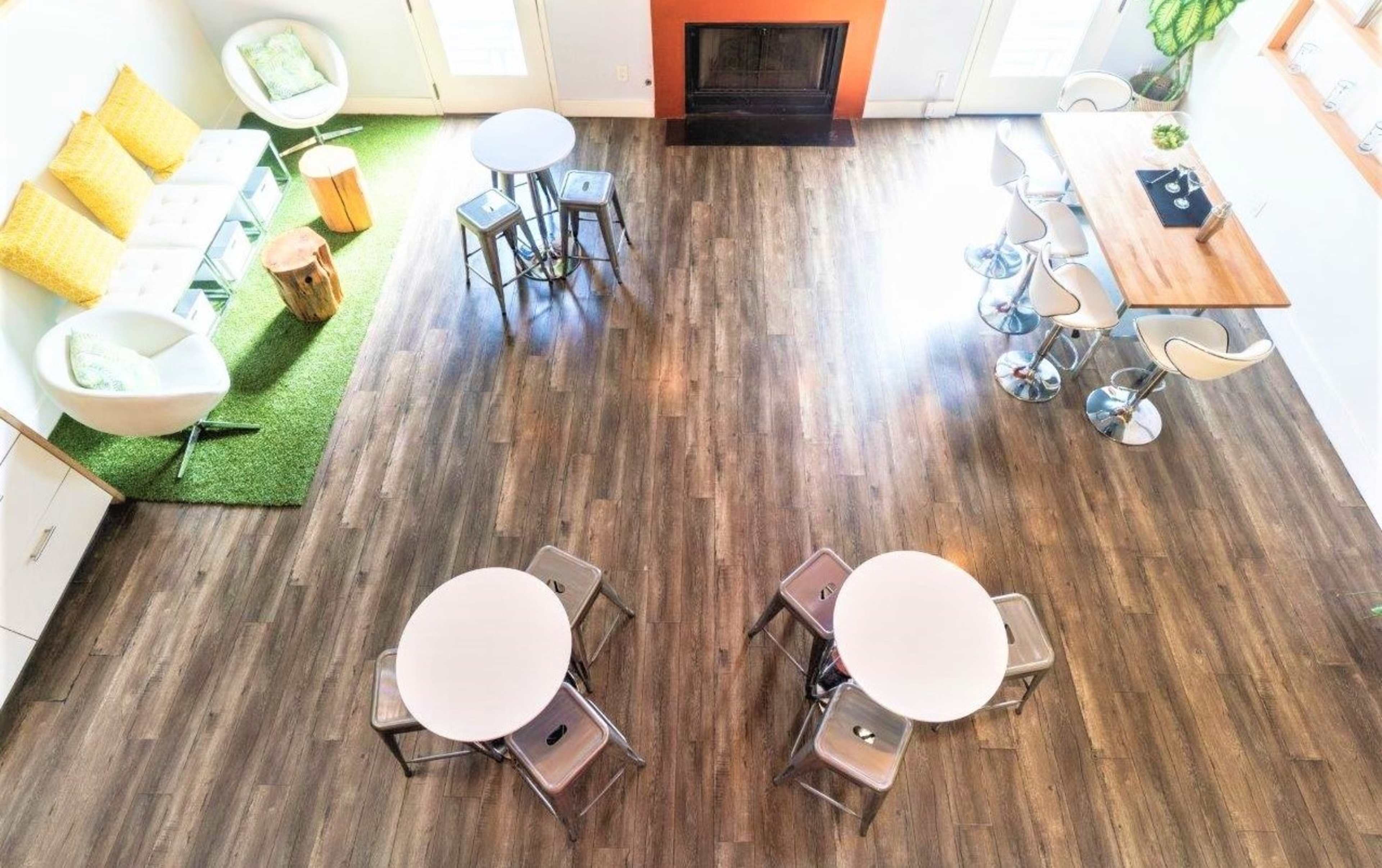 The image shows a spacious room with wooden flooring, featuring two circular tables surrounded by metal chairs, a sitting area with a white couch and yellow cushions, and a bar-style table with stools near a fireplace.