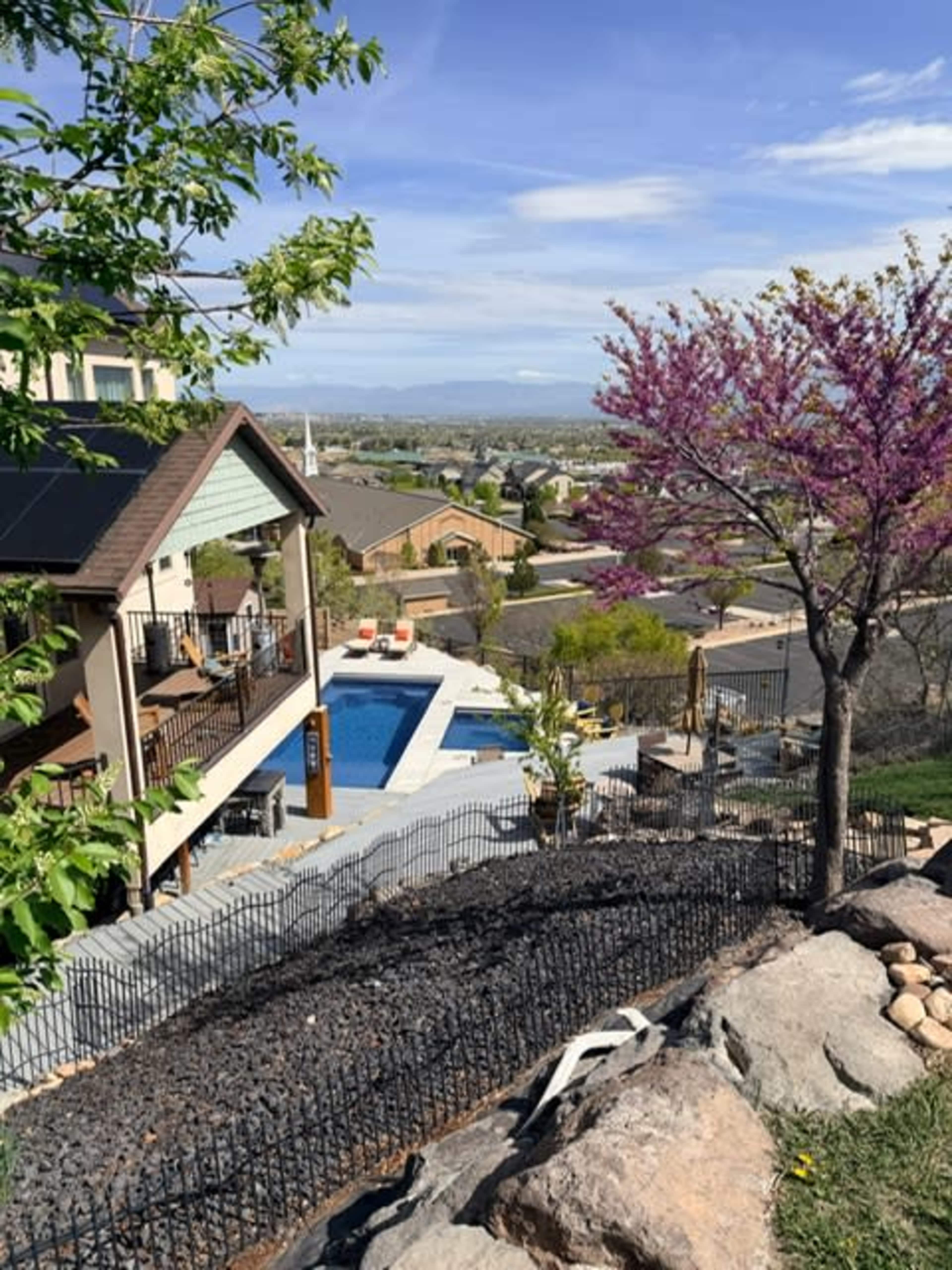 The image shows a hillside residential property with a swimming pool, surrounded by landscaped areas, and overlooking a view of distant houses and hills.