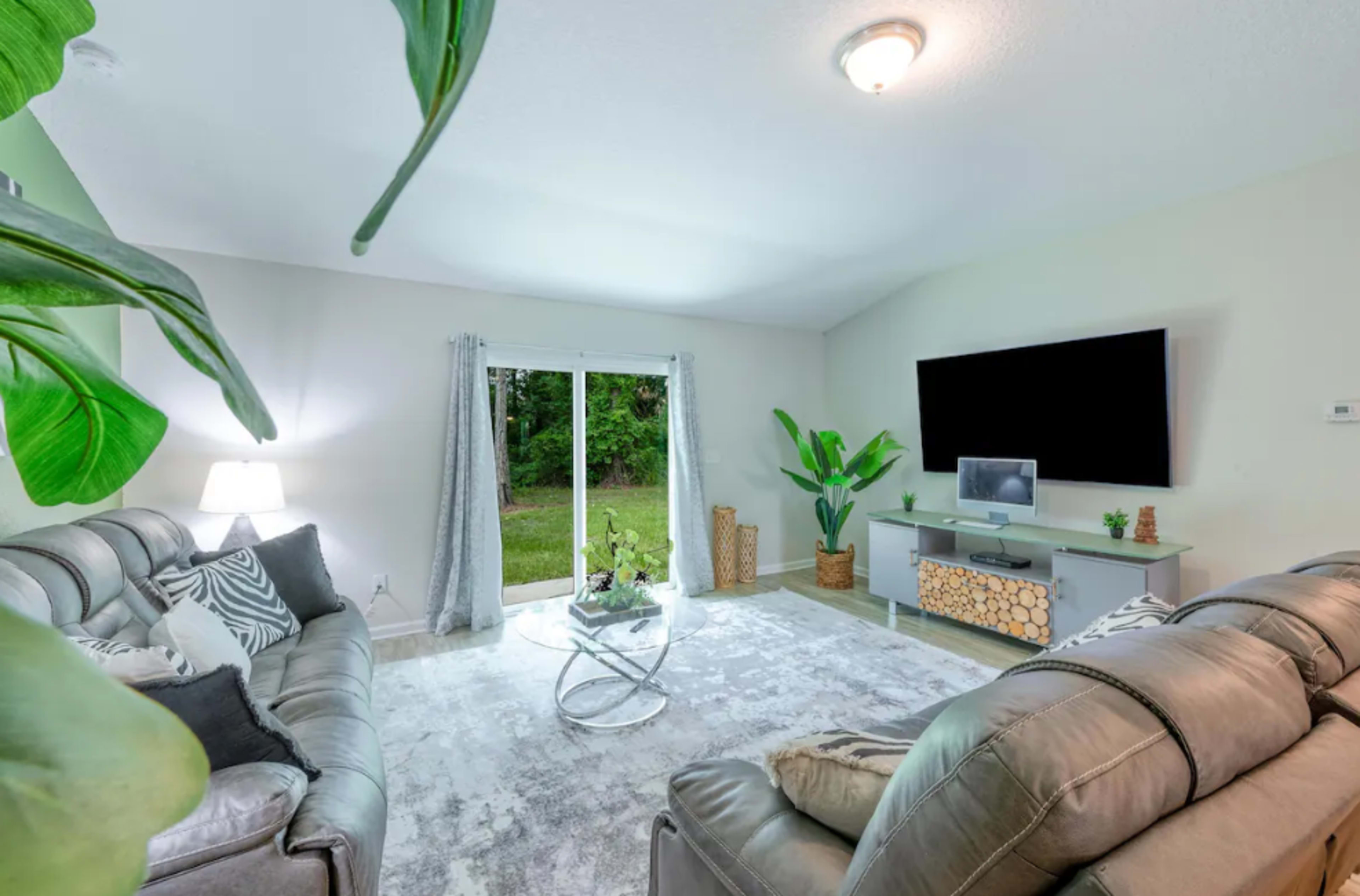 A modern living room with two gray sofas, a glass coffee table, a television mounted on the wall, and large windows overlooking a green landscape.