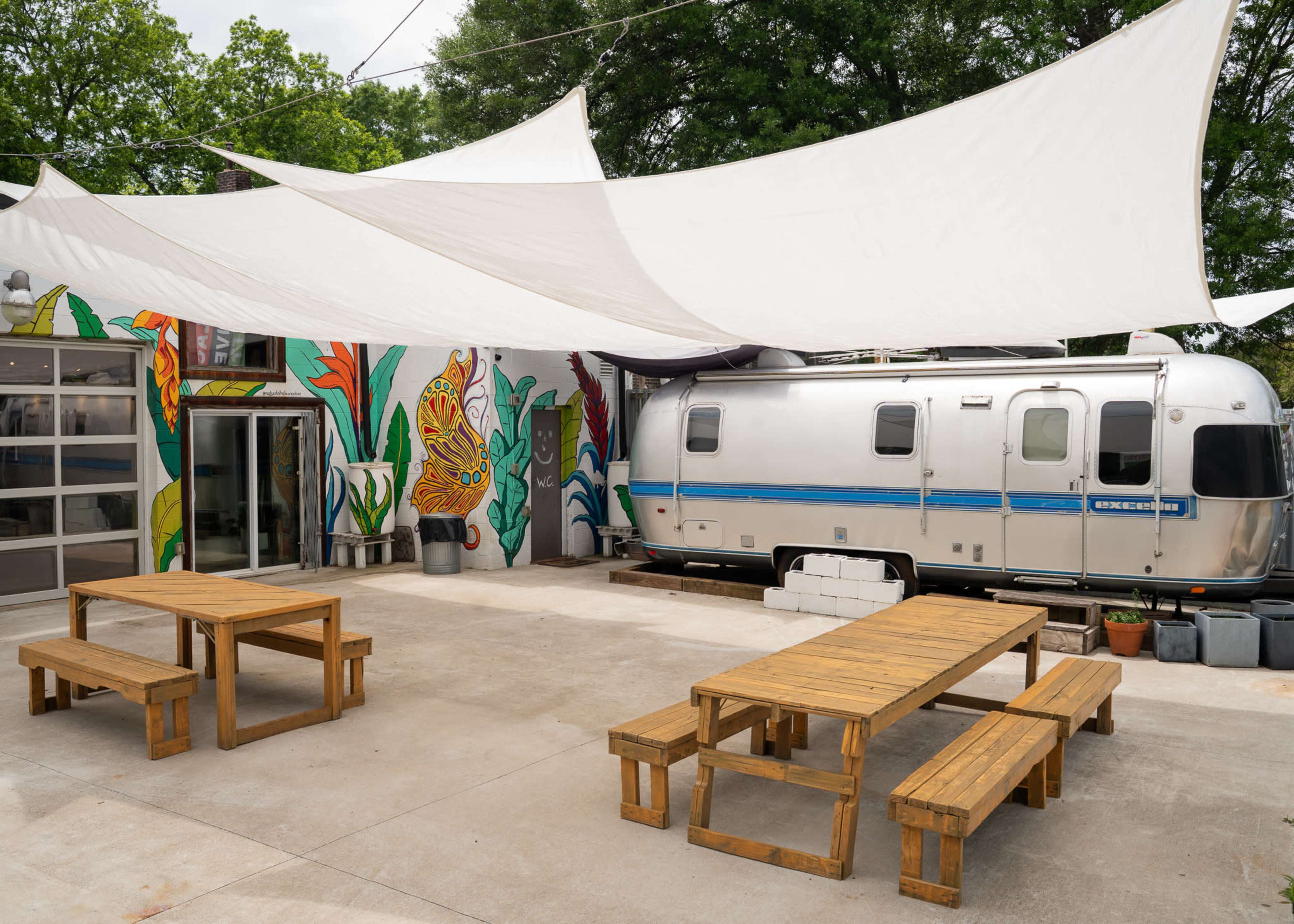 The image shows an outdoor area with wooden picnic tables under a large shade structure near a silver Airstream trailer and a colorful mural.