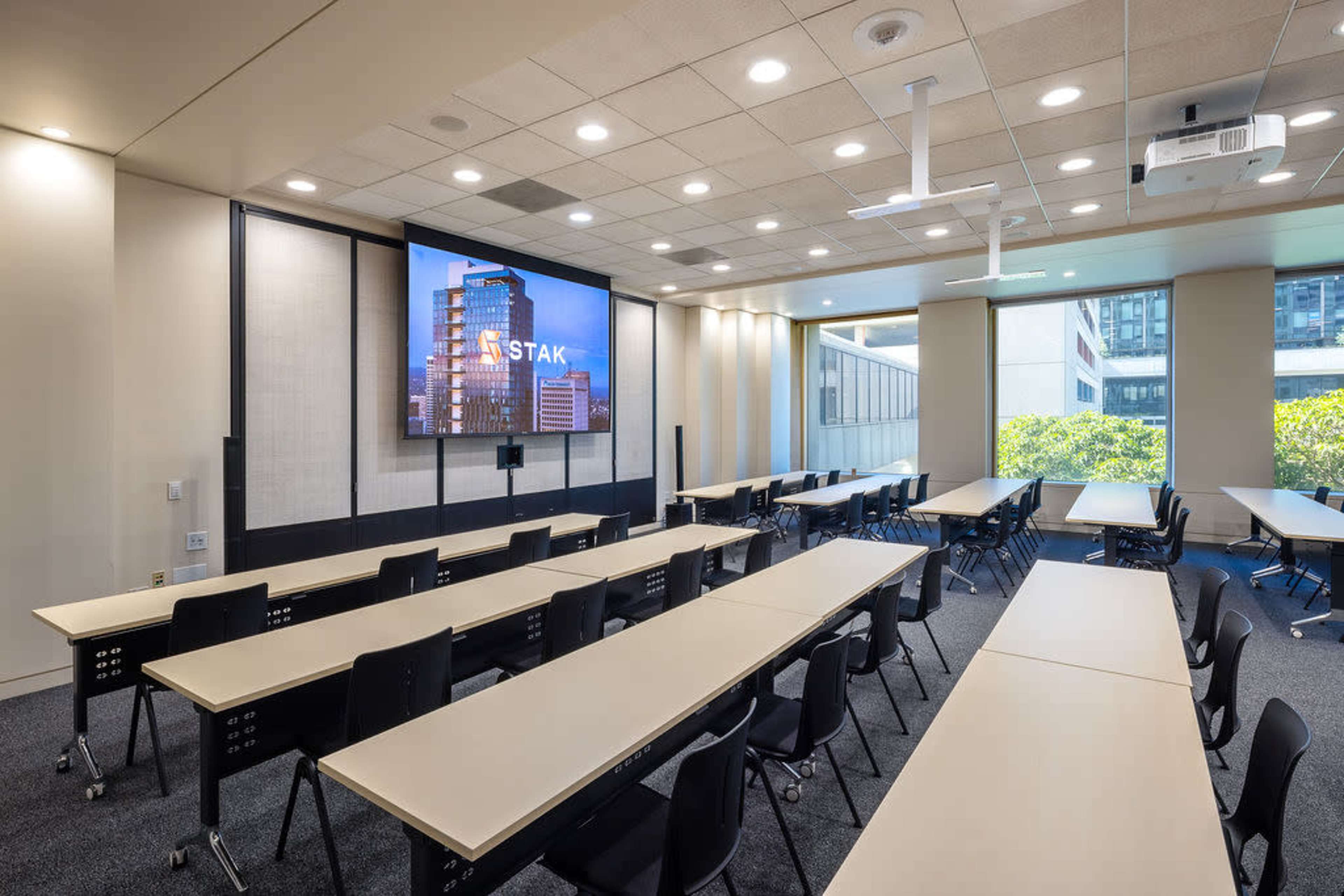 The image shows a modern conference room furnished with tables and chairs, featuring a large screen displaying the word "STAK" on one wall.