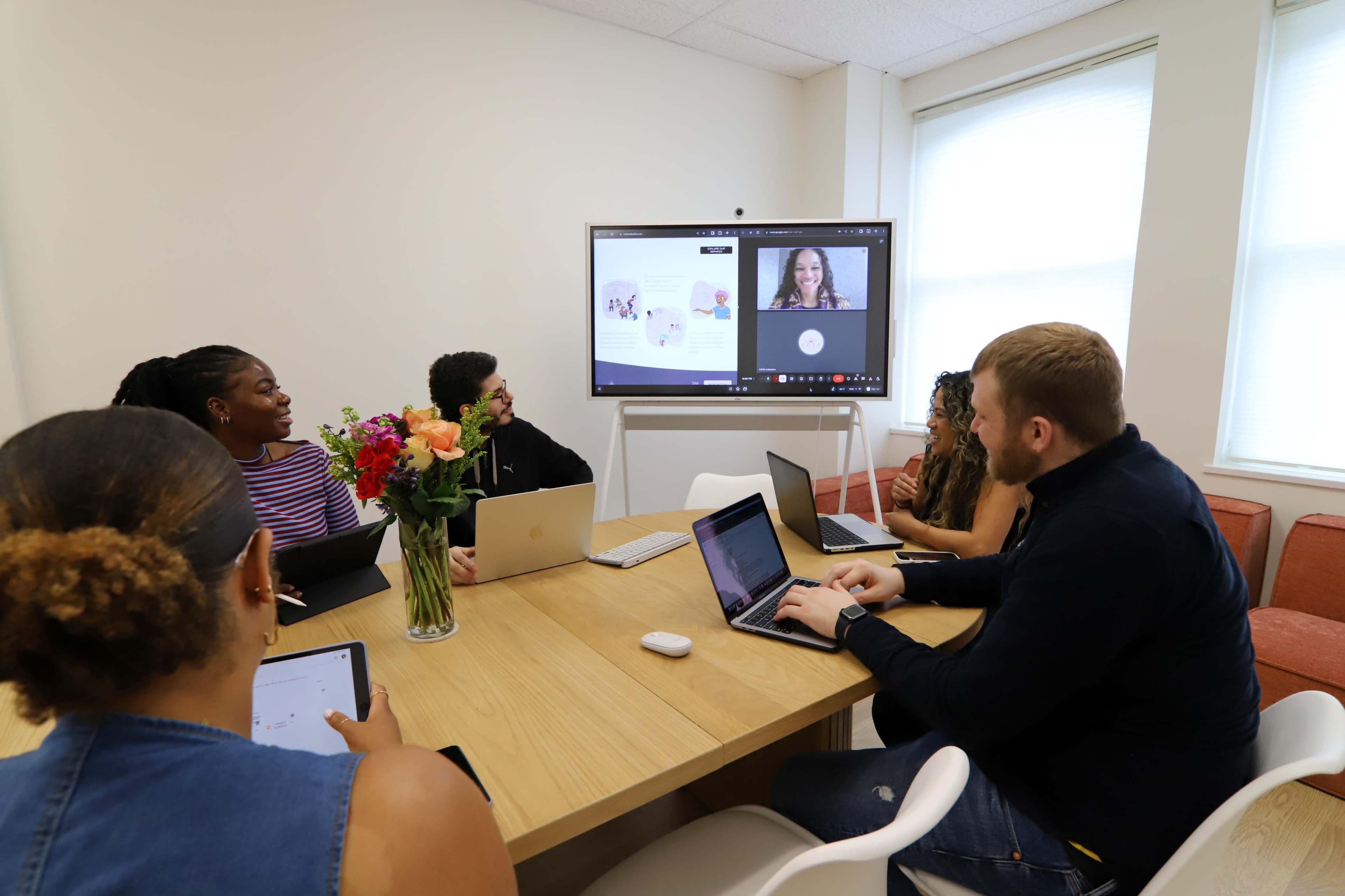 A group of six people are sitting around a wooden table in a modern meeting room, collaborating while using laptops and a large screen displays a video call.
