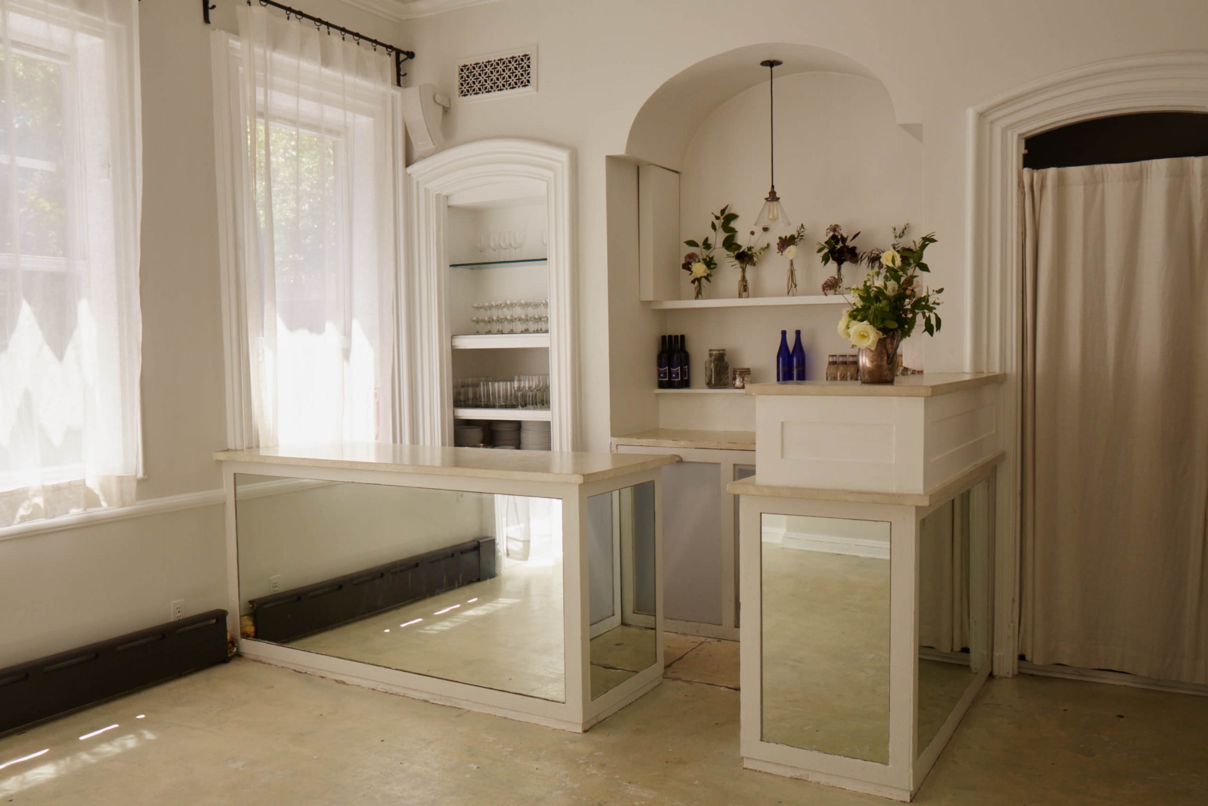 The image shows a bright room with large windows, featuring a bar area with a mirrored counter and shelves displaying bottles and a floral arrangement.