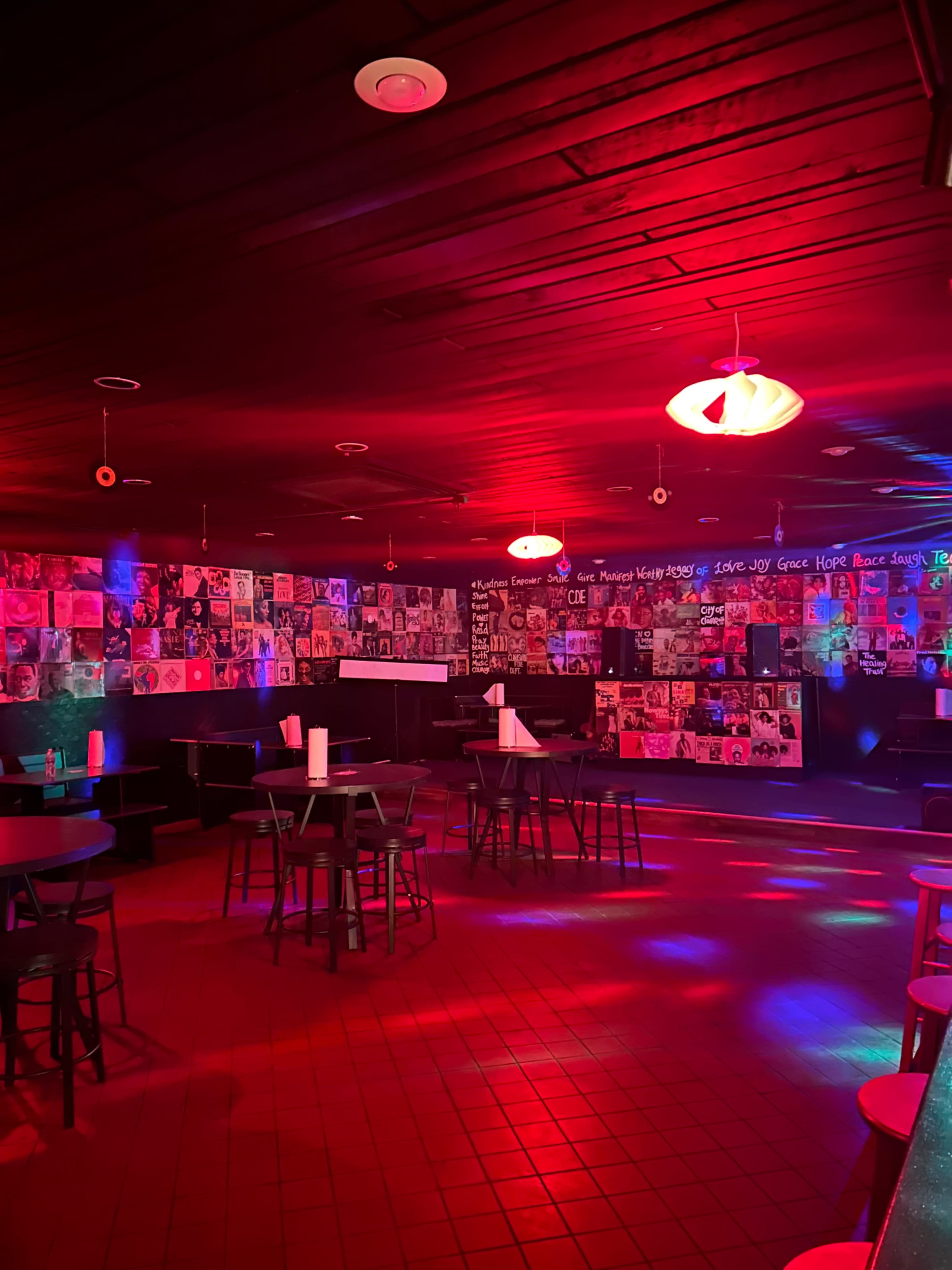 The scene shows a dimly lit café with red and blue lighting, featuring tables and a wall covered in posters and photographs.