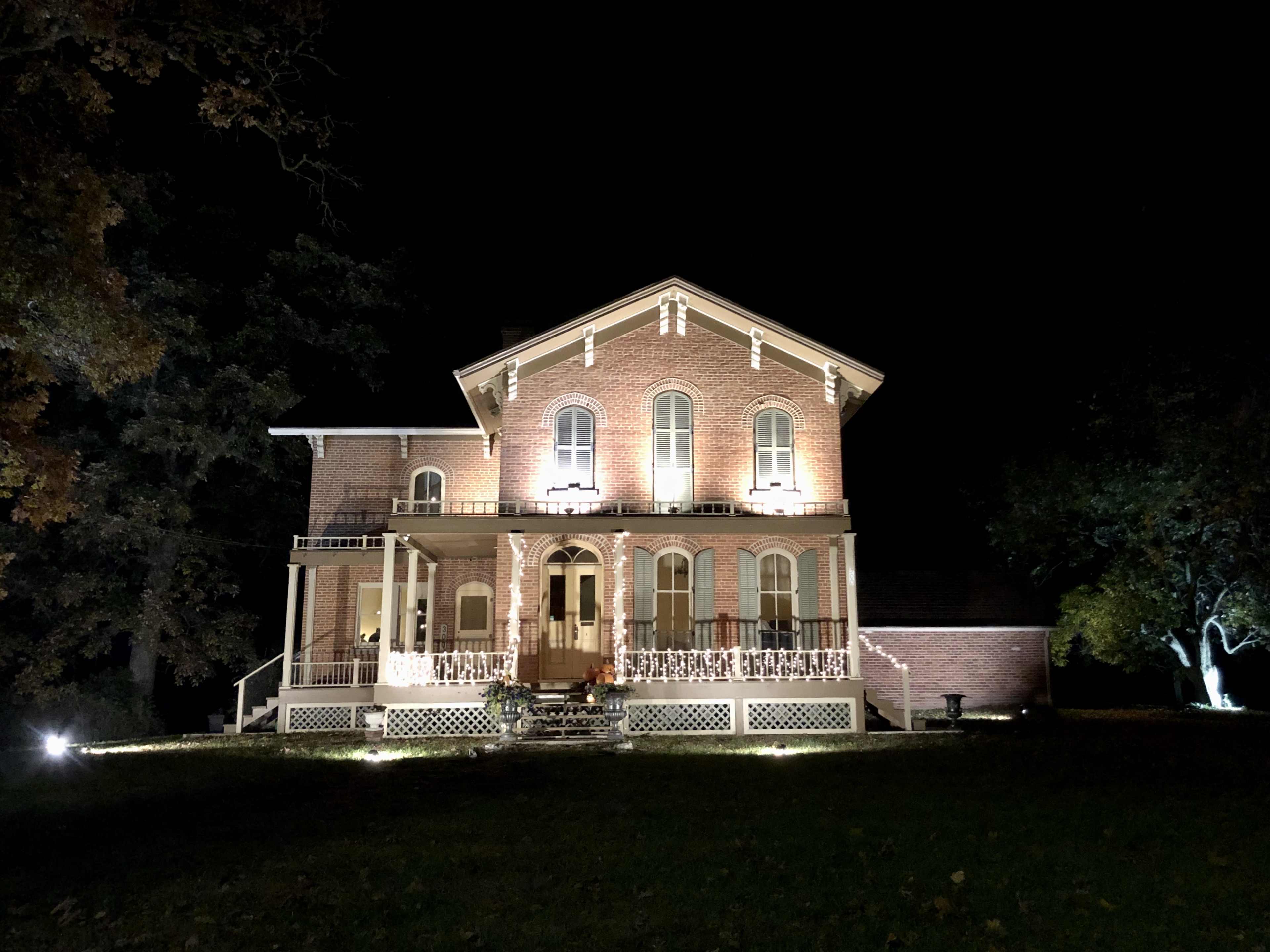 A brick house with a prominent porch is illuminated at night, surrounded by trees and a well-maintained lawn.