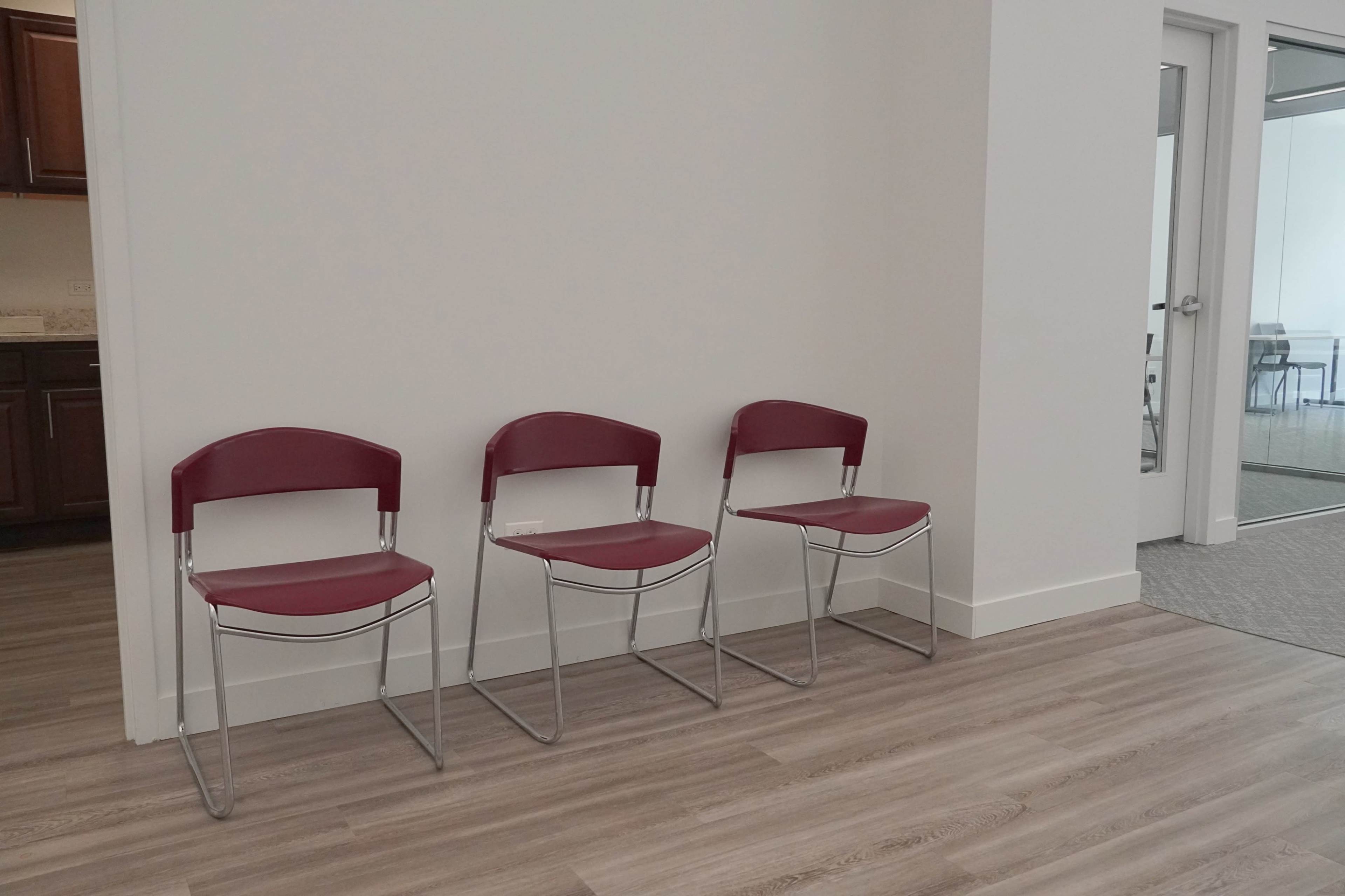 Three red folding chairs are aligned against a light-colored wall in a room with a wooden floor.
