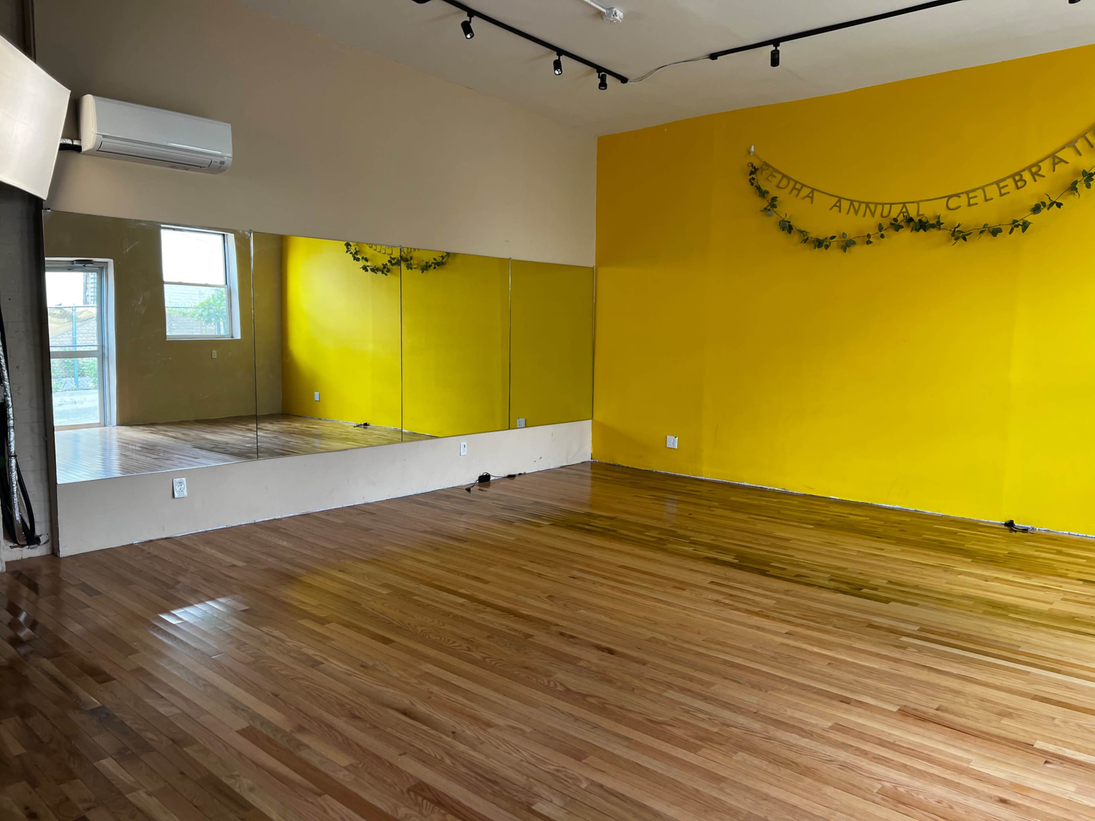 The image shows a spacious studio with yellow walls, a wooden floor, and a large mirror along one side.