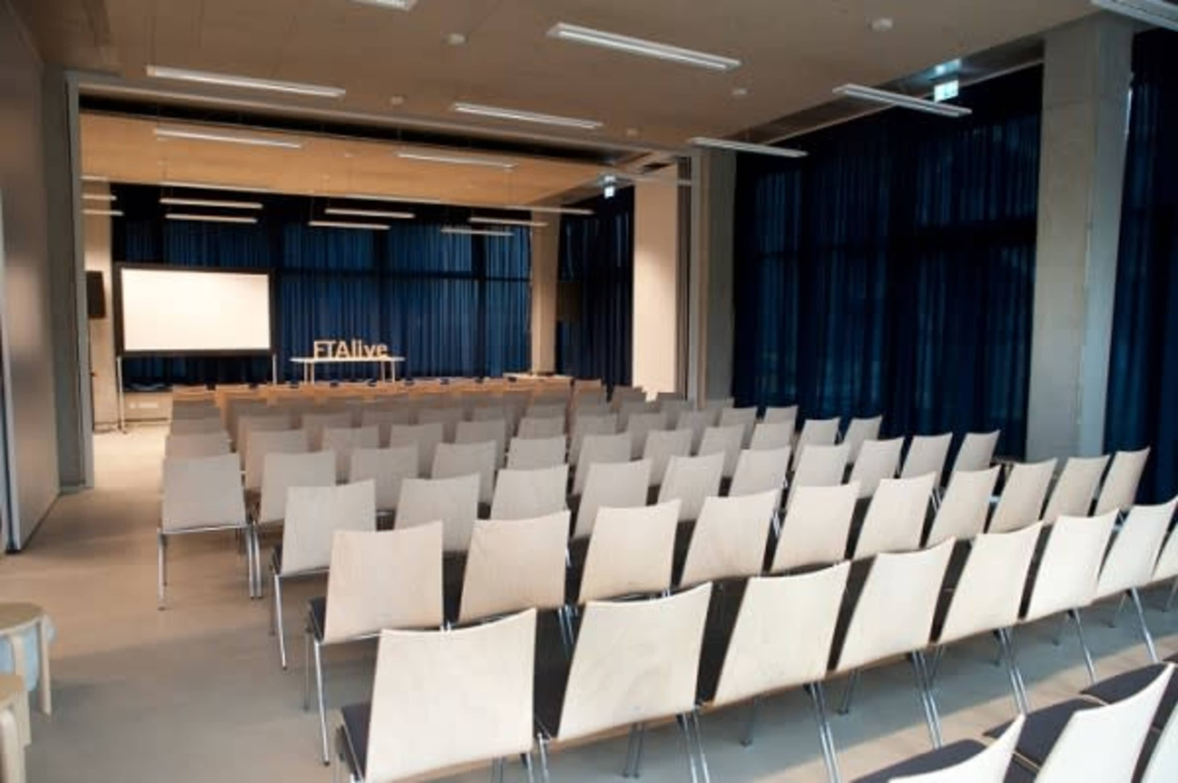 The image shows a spacious conference room with rows of empty wooden chairs facing a large screen and a backdrop with the text "#Alive."