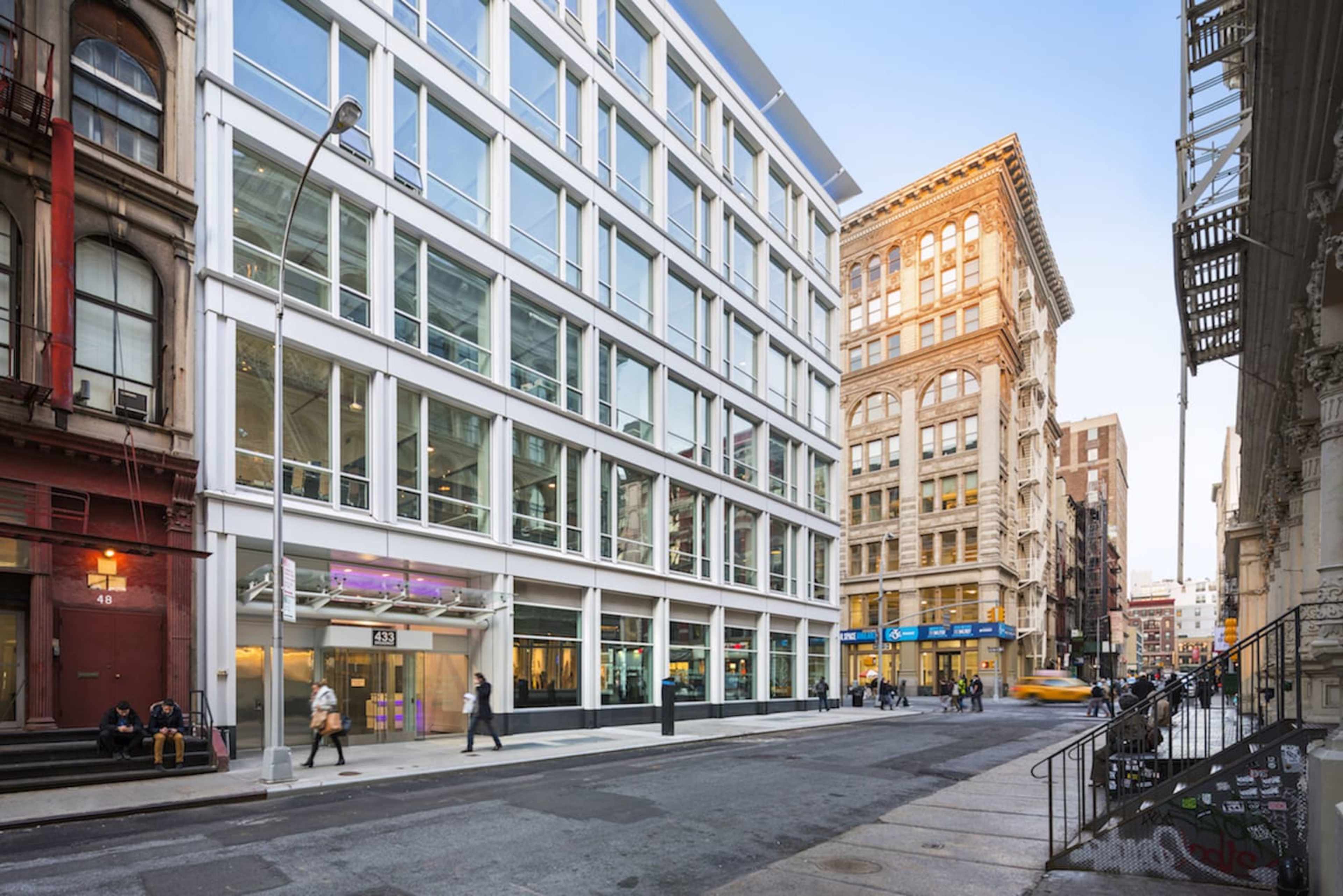 The image shows a modern glass building at the corner of a street, adjacent to older brick buildings in an urban setting.