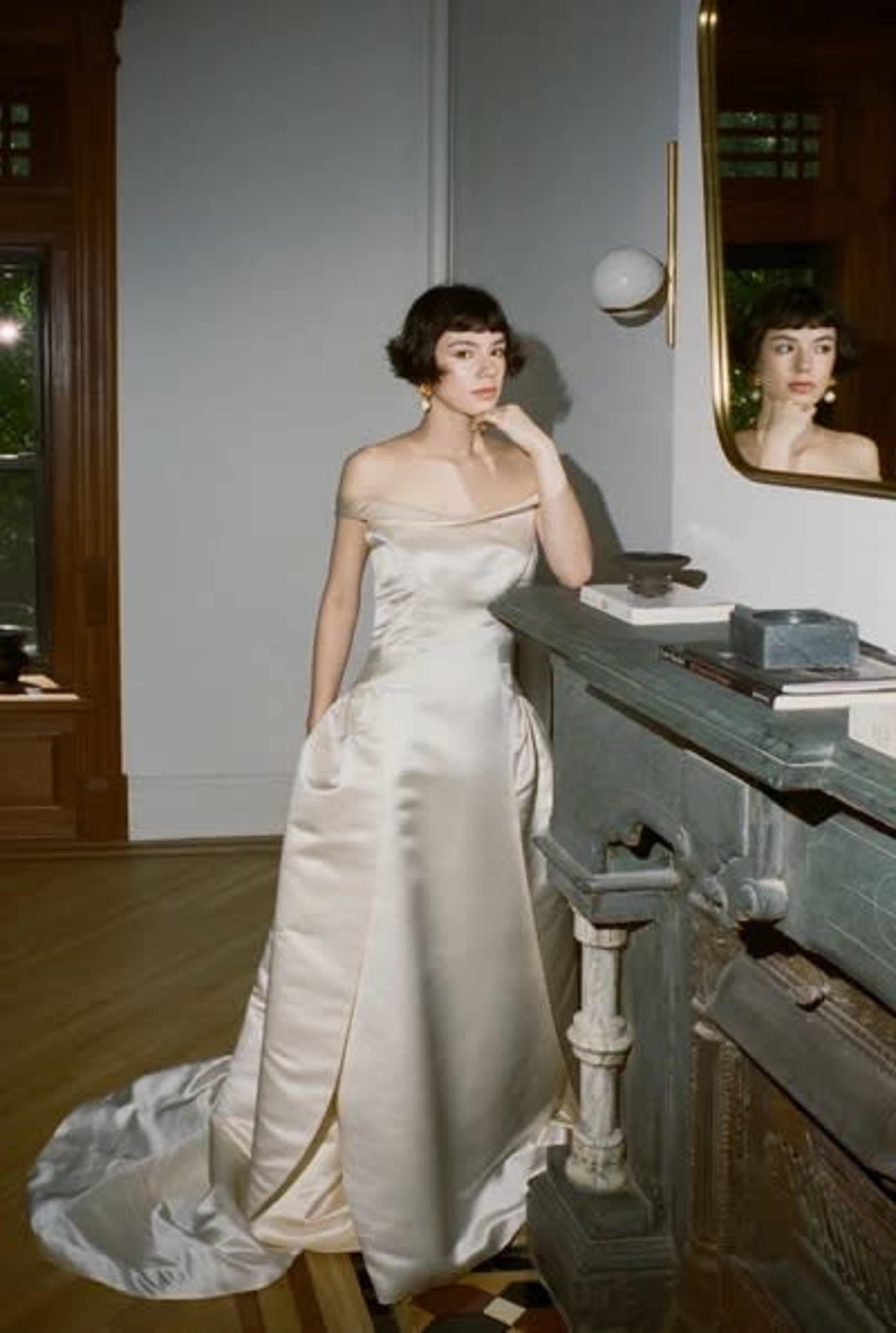 A woman in a satin off-shoulder gown stands next to a fireplace, gazing thoughtfully at her reflection in a mirror.