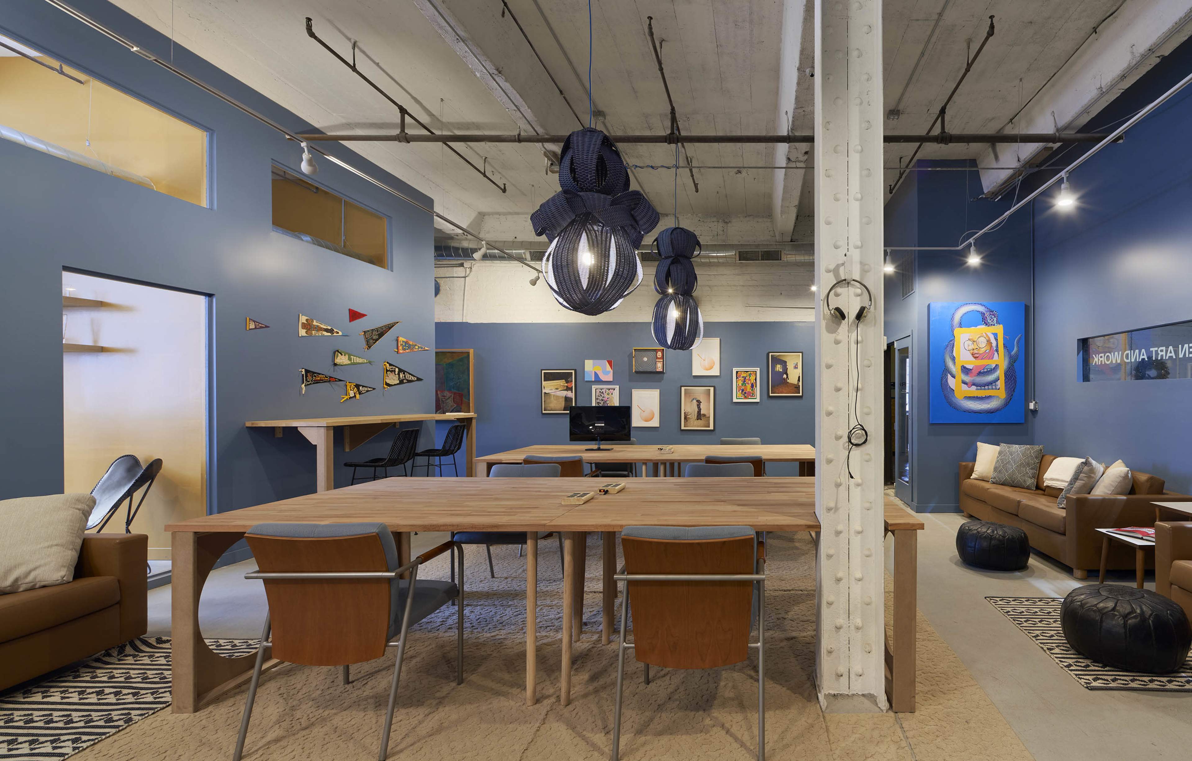 The image shows a spacious open-plan office featuring wooden tables, modern pendant lighting, and colorful wall art.