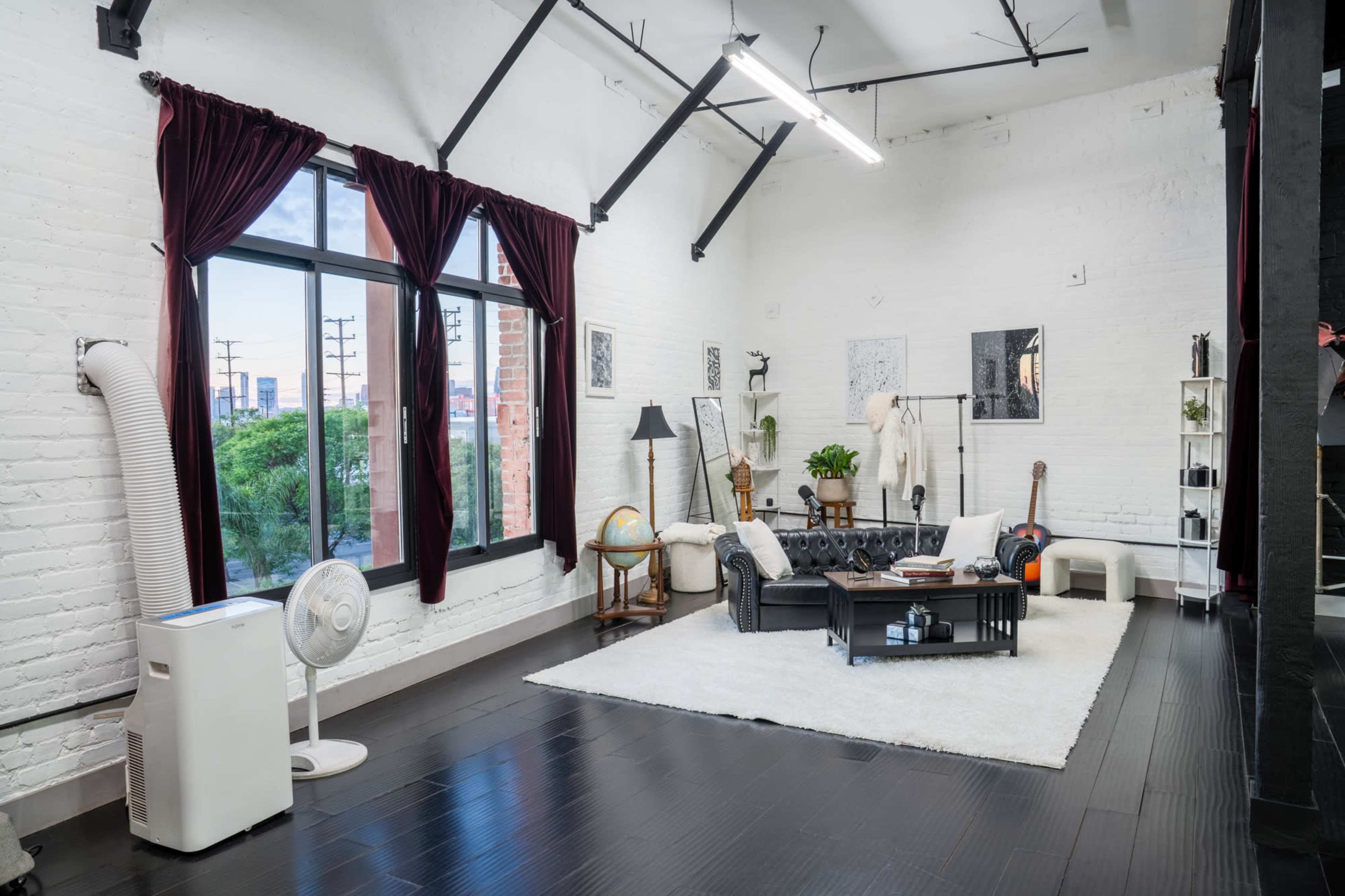 The image shows a modern, bright living space with a black leather sofa, a coffee table, a globe, and various decorative items placed on a white rug beneath large windows with dark red curtains.