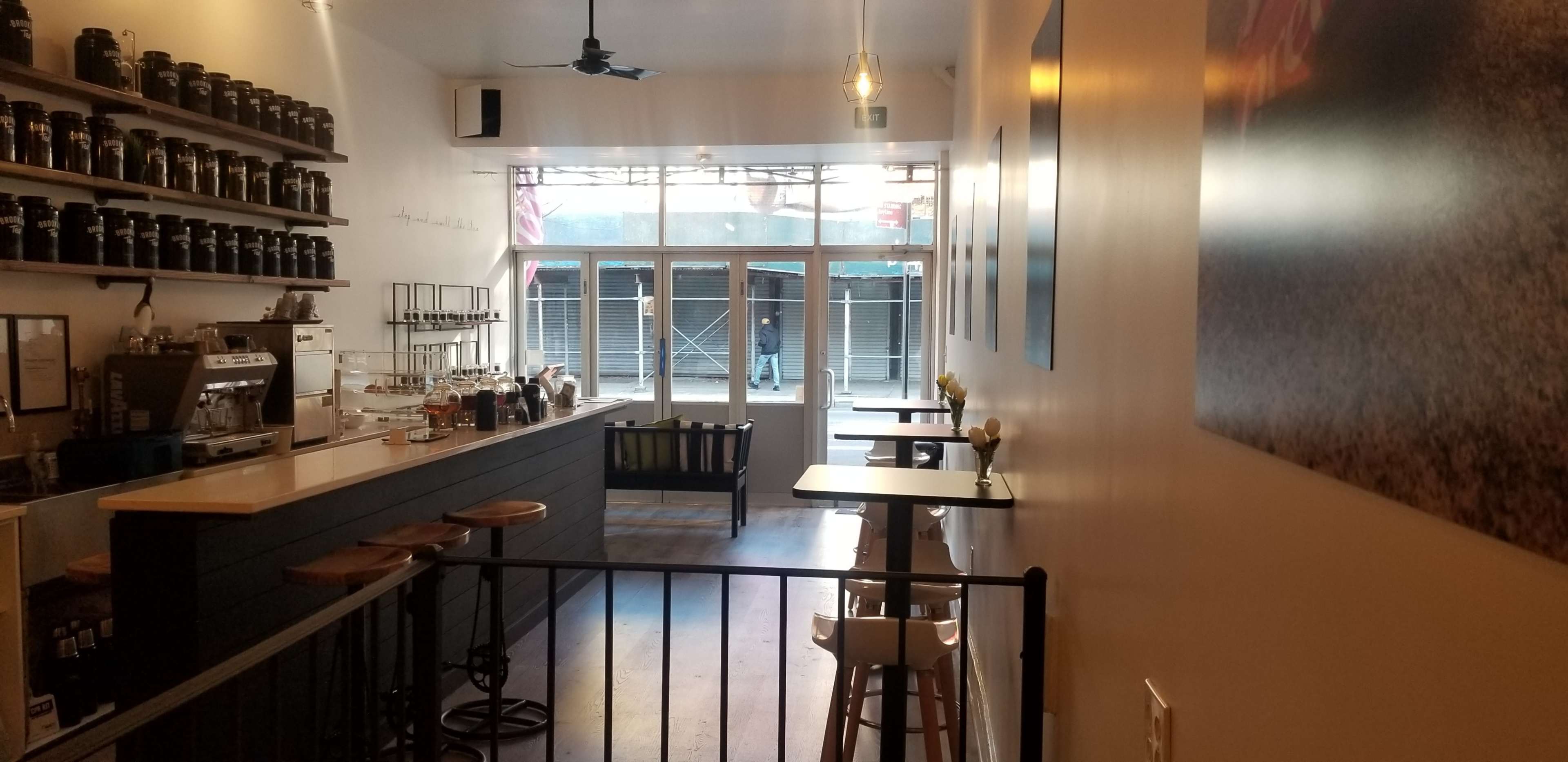 The image shows a coffee shop interior with a counter on the left, bar stools, and tables, leading to a large window facing the street.