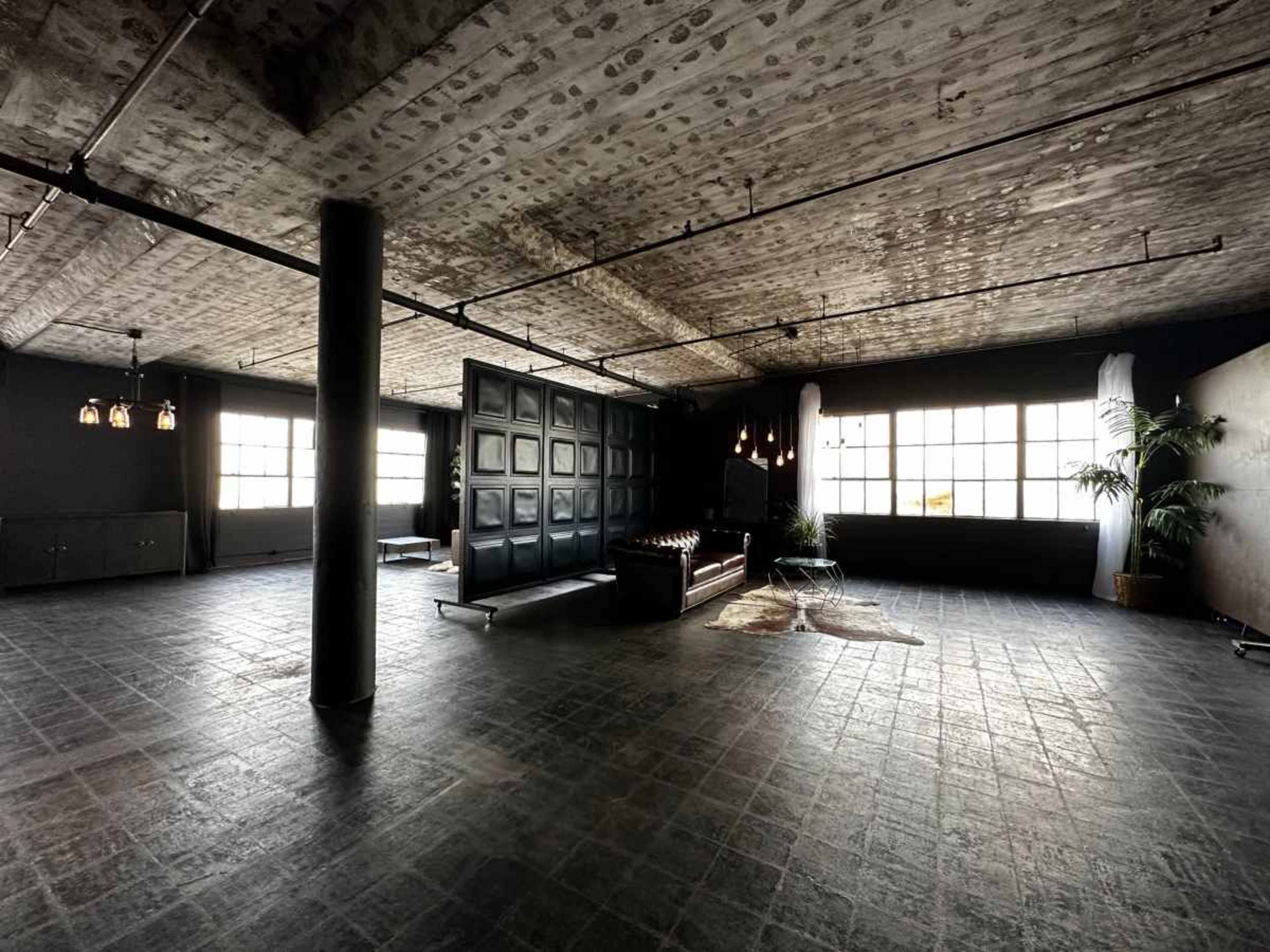 The image shows a spacious industrial-style interior with large windows, a dark floor, and minimalist furniture including a leather sofa and a decorative rug.
