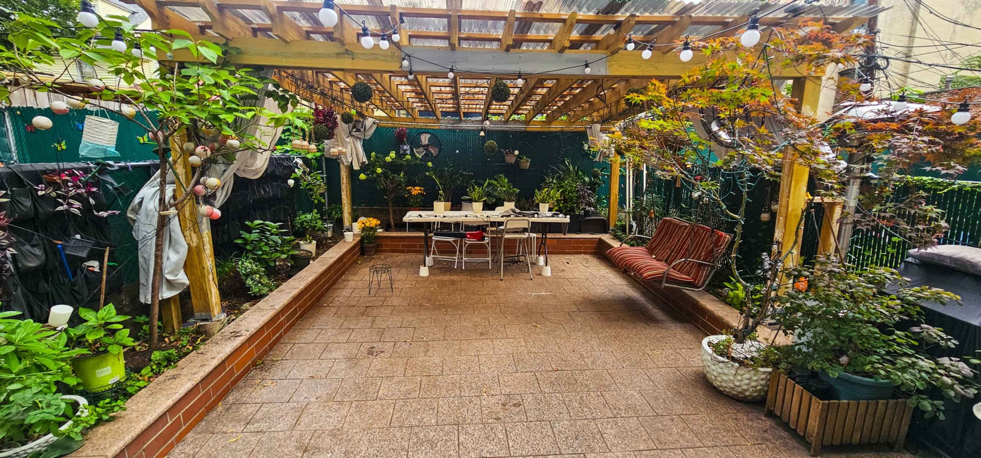 The image shows a landscaped patio area with a wooden pergola, a dining table, chairs, and various potted plants arranged around the space.