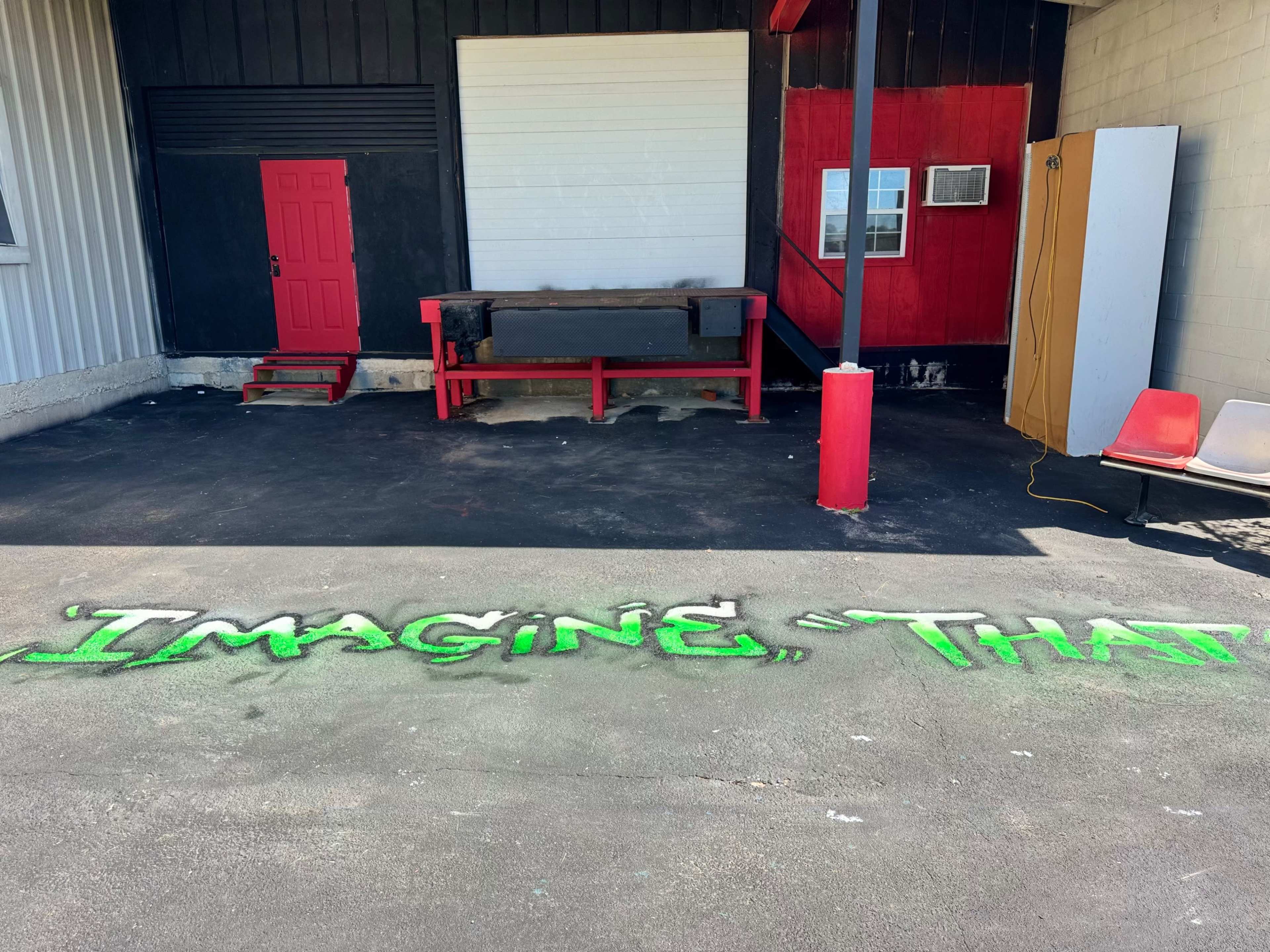 The image shows a partially enclosed outdoor area with a red door, a red bench, and the phrase "Imagine, That" painted in green on the ground.