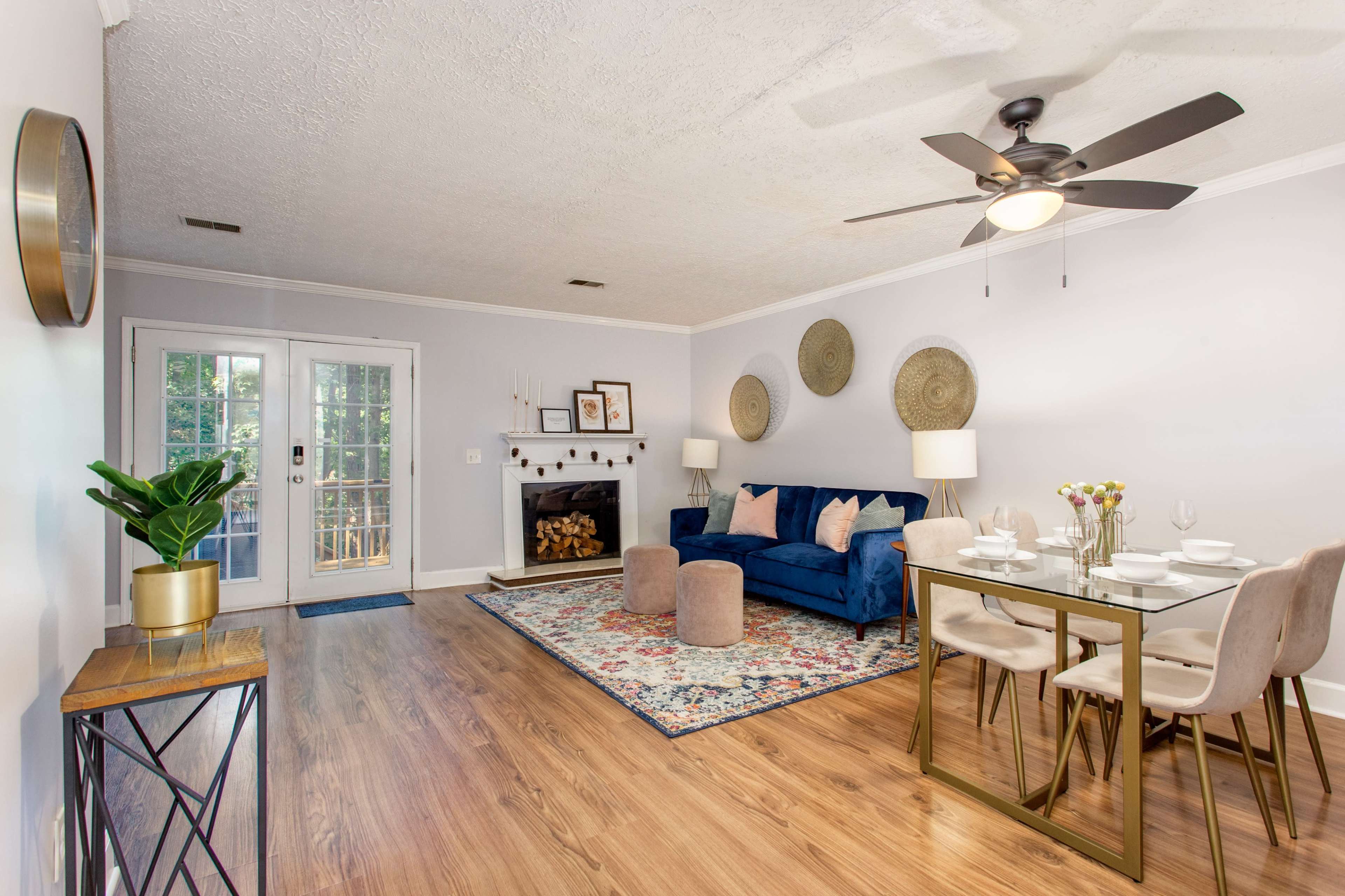 A living room with a blue sofa, a glass dining table set for four, and a fireplace, alongside large French doors leading to an outdoor area.